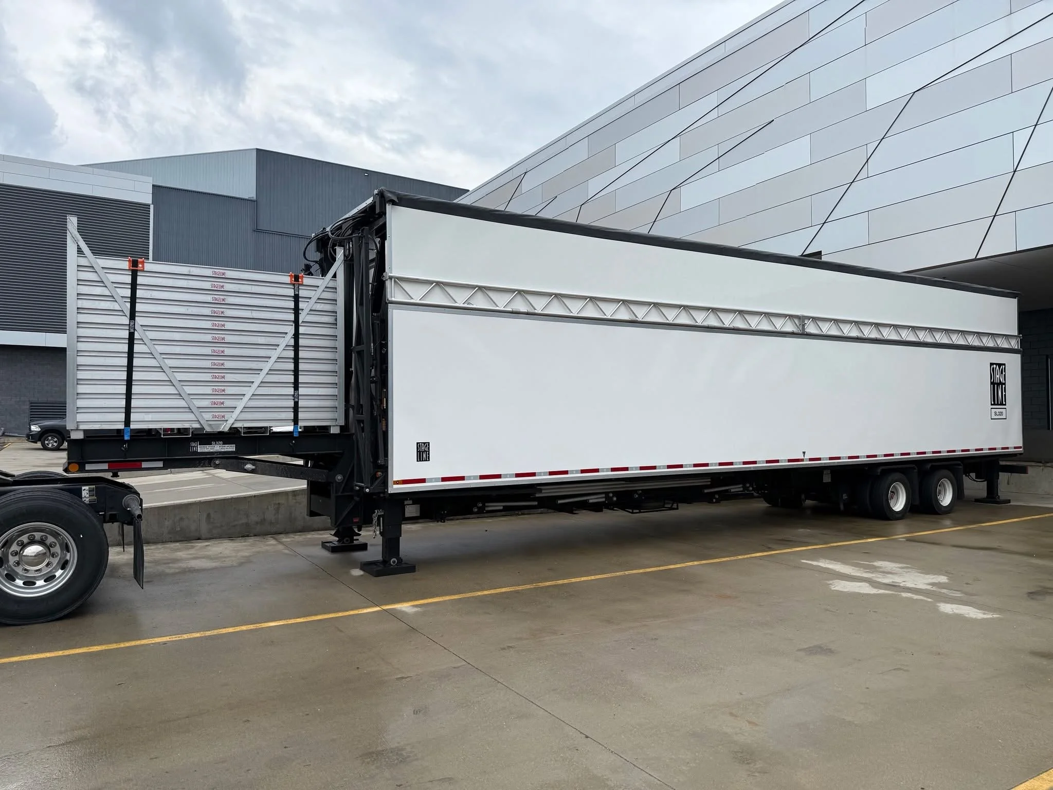 A large truck trailer parked in a parking lot with a modern building in the background. The trailer is white with a black and white logo and is partially loaded or empty. The parking lot has marked spaces and some cars are visible in the distance.