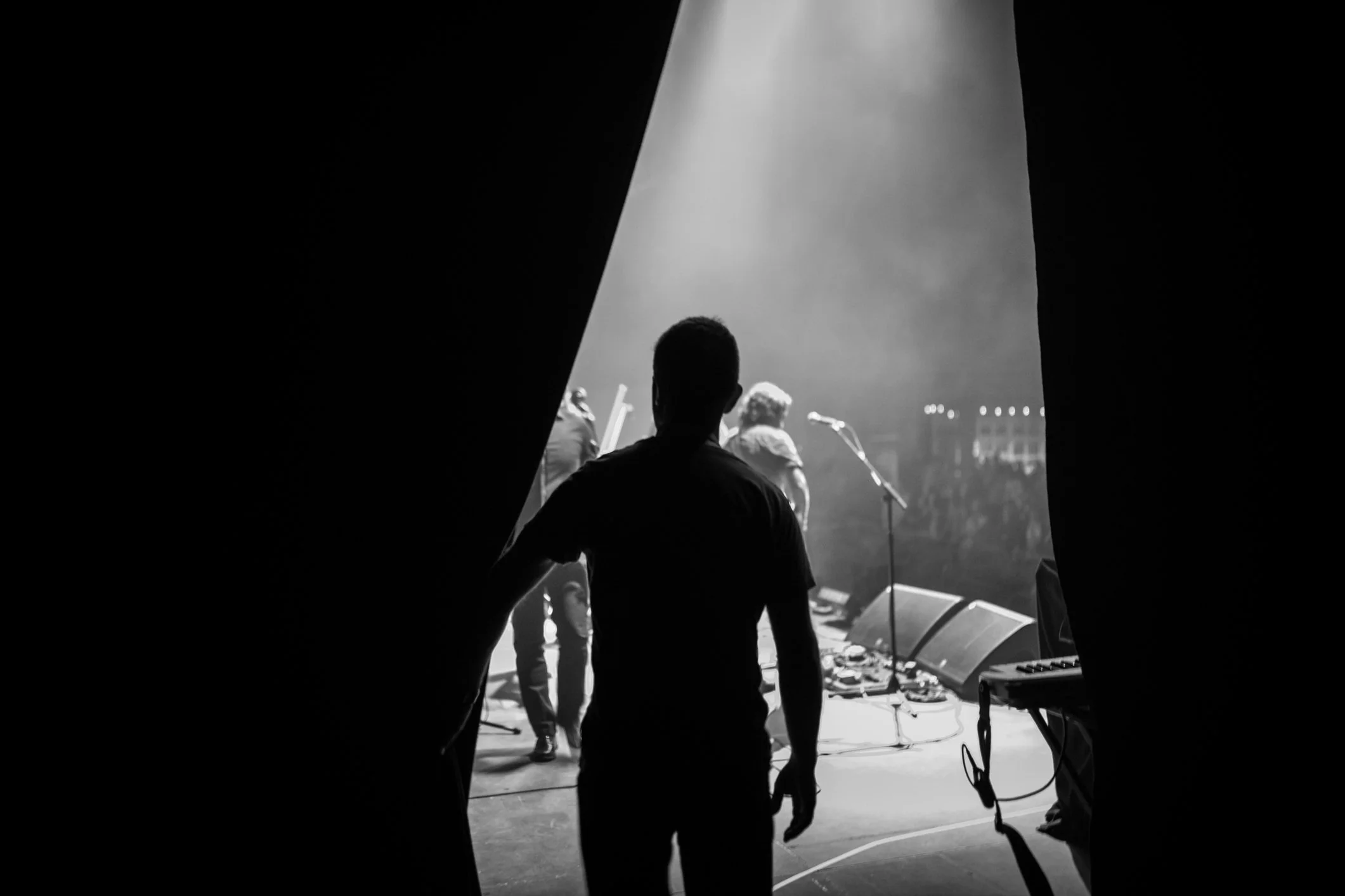 Black and white photo of a man watching a band perform on stage through a partially opened curtain.