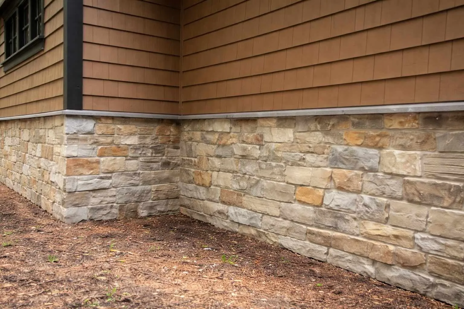 The corner of a house showing a layered stone foundation and wood siding above it, with a small amount of soil and some small plants at the base.