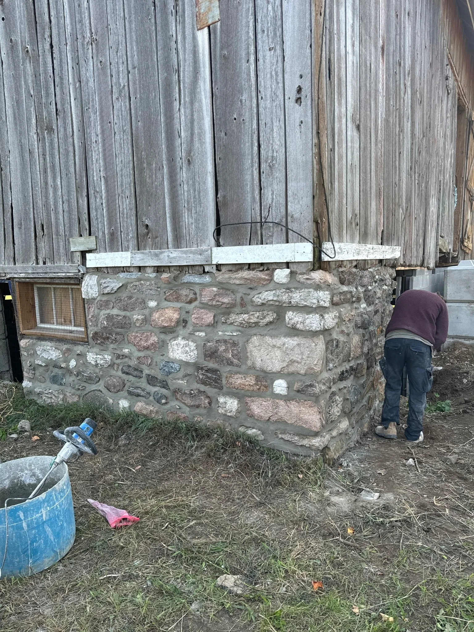 Person working on stone foundation with wooded exterior wall above, construction tools and materials nearby.