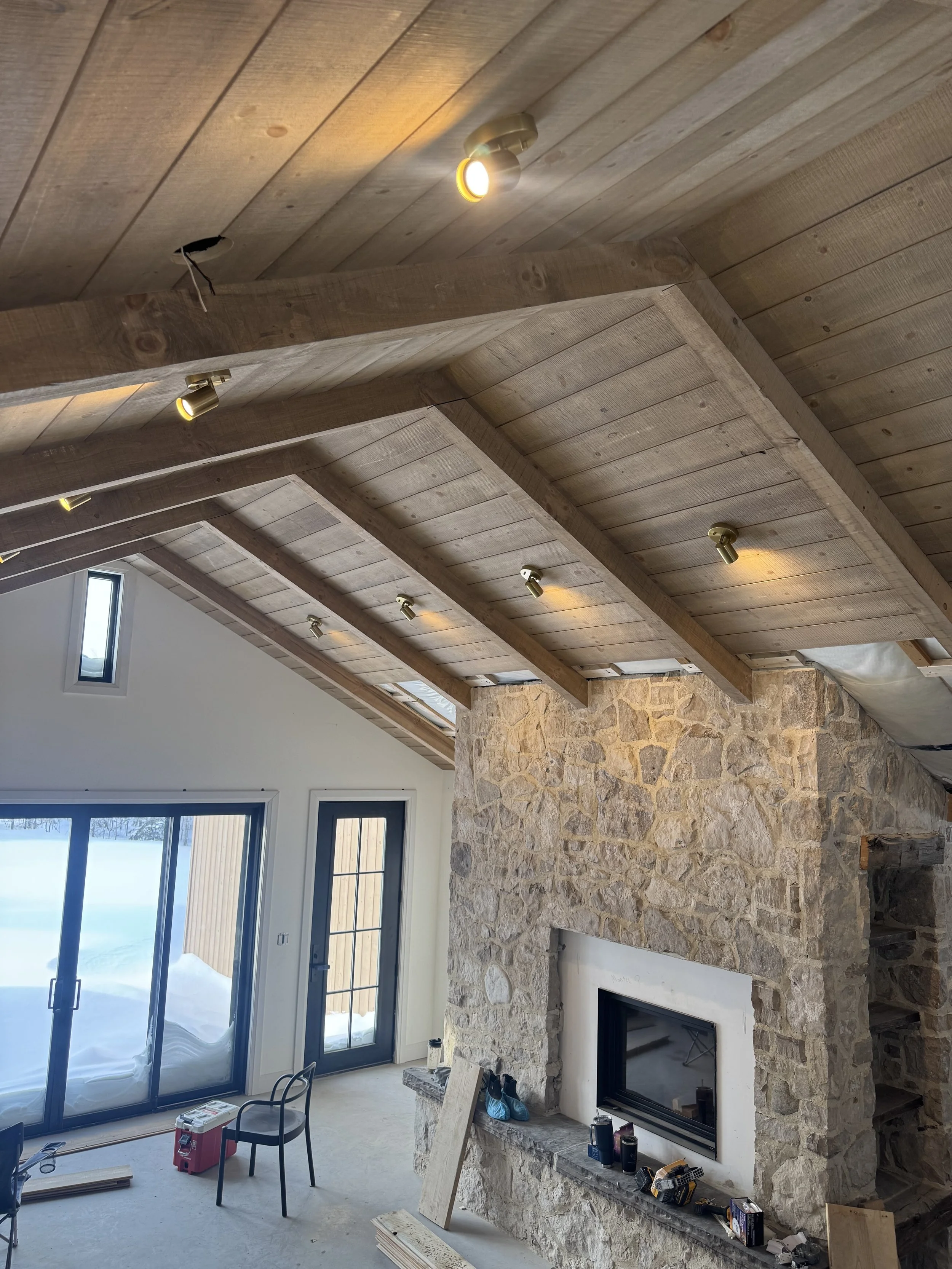 Interior view of a living room with a sloped wooden ceiling featuring multiple small profile lights, a stone fireplace with tools and materials nearby, a sliding glass door and a smaller window showing snow outside, and various construction tools and supplies on the floor and ledges.