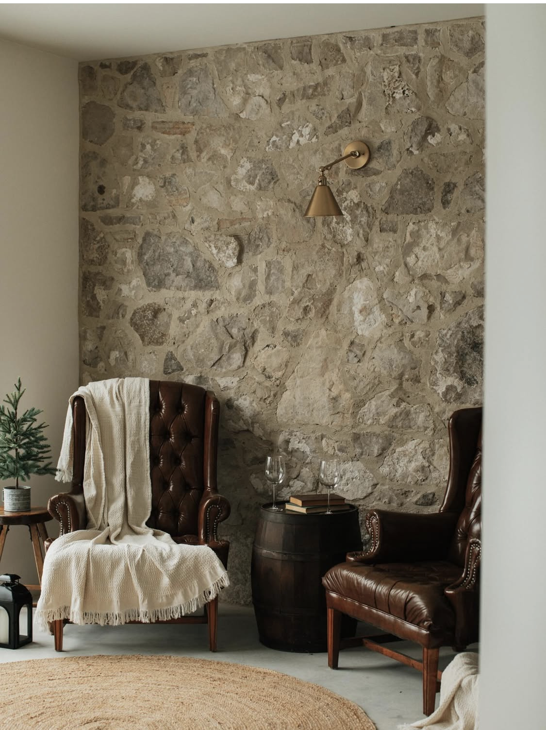 A cozy living room corner with a stone accent wall, two brown leather armchairs, a small potted pine tree on a wooden side table, a round wooden barrel table with two wine glasses and books, a beige throw blanket draped over one armchair, a black lantern on the floor, a round woven rug on the light-colored floor, and a gold adjustable wall-mounted reading lamp.