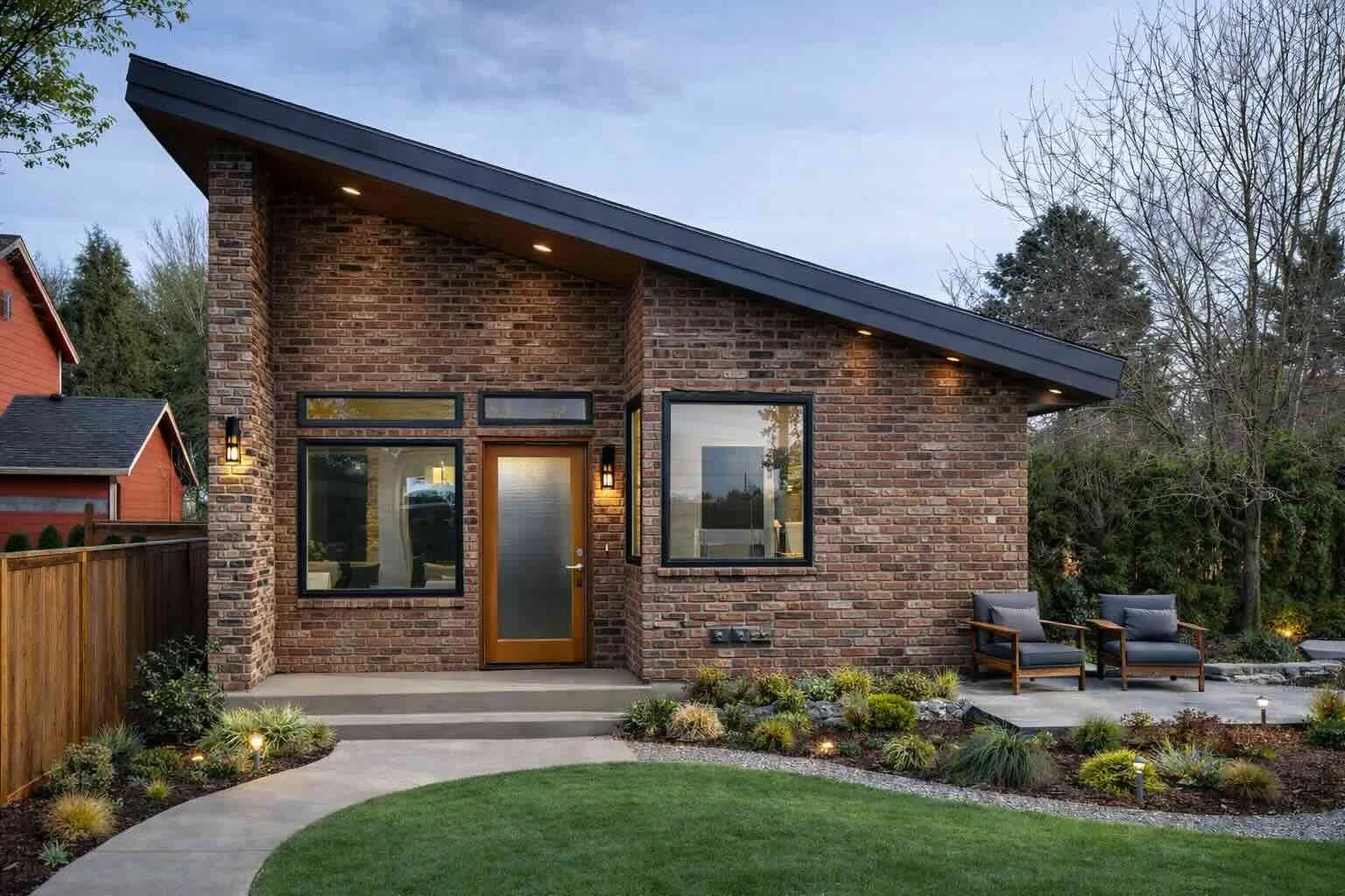 Modern brick house with large windows, a sloped roof with outdoor lighting, sitting area with two chairs, a manicured lawn, and landscaped garden with small plants and pathway.