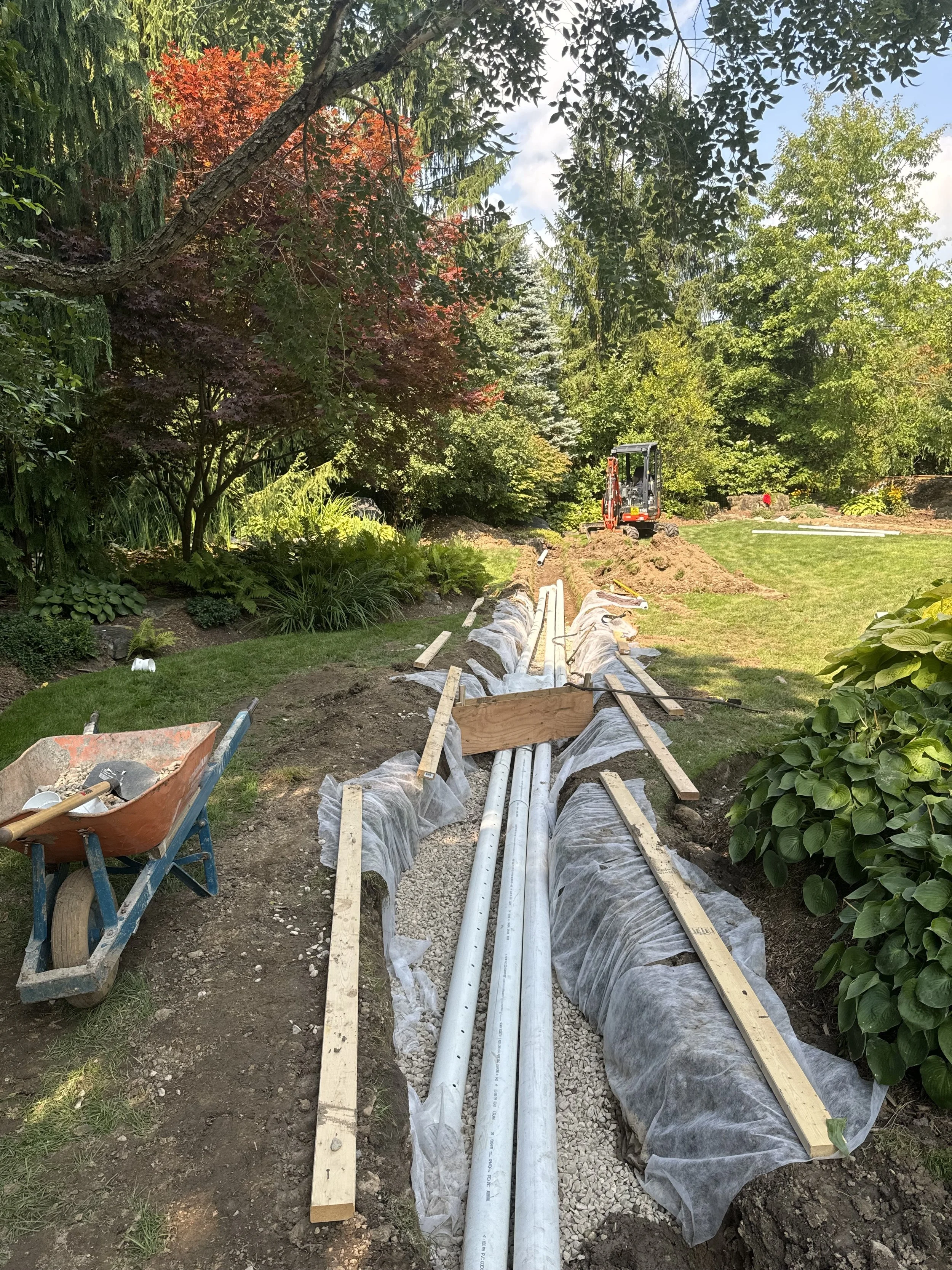 Garden landscaping project with underground pipes being installed, a wheelbarrow filled with tools on the left, and a small excavator in the background amidst trees and plants.