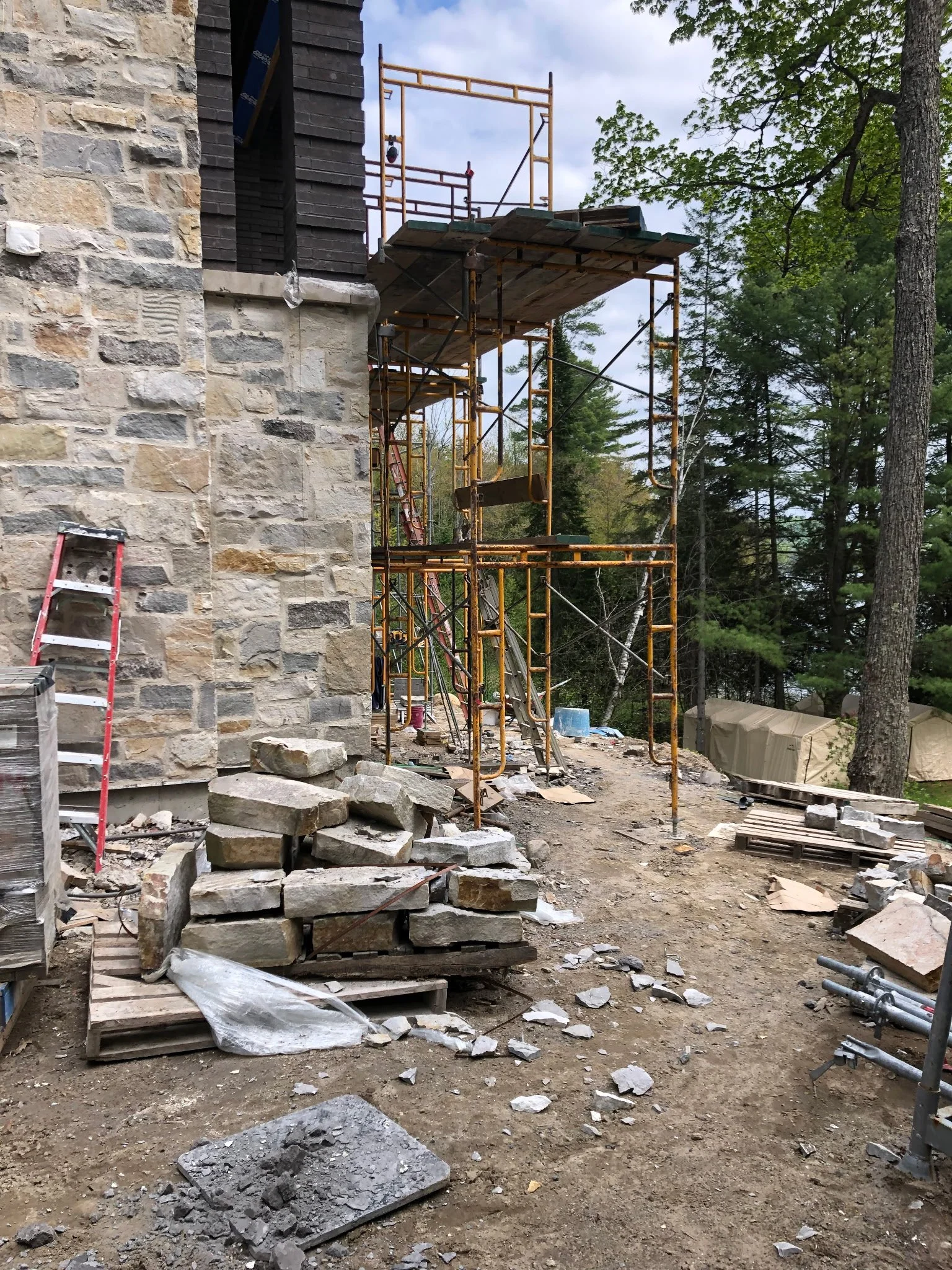 Construction site with scaffolding next to a stone building, scattered rocks, ladders, and construction materials amidst trees and outdoors.
