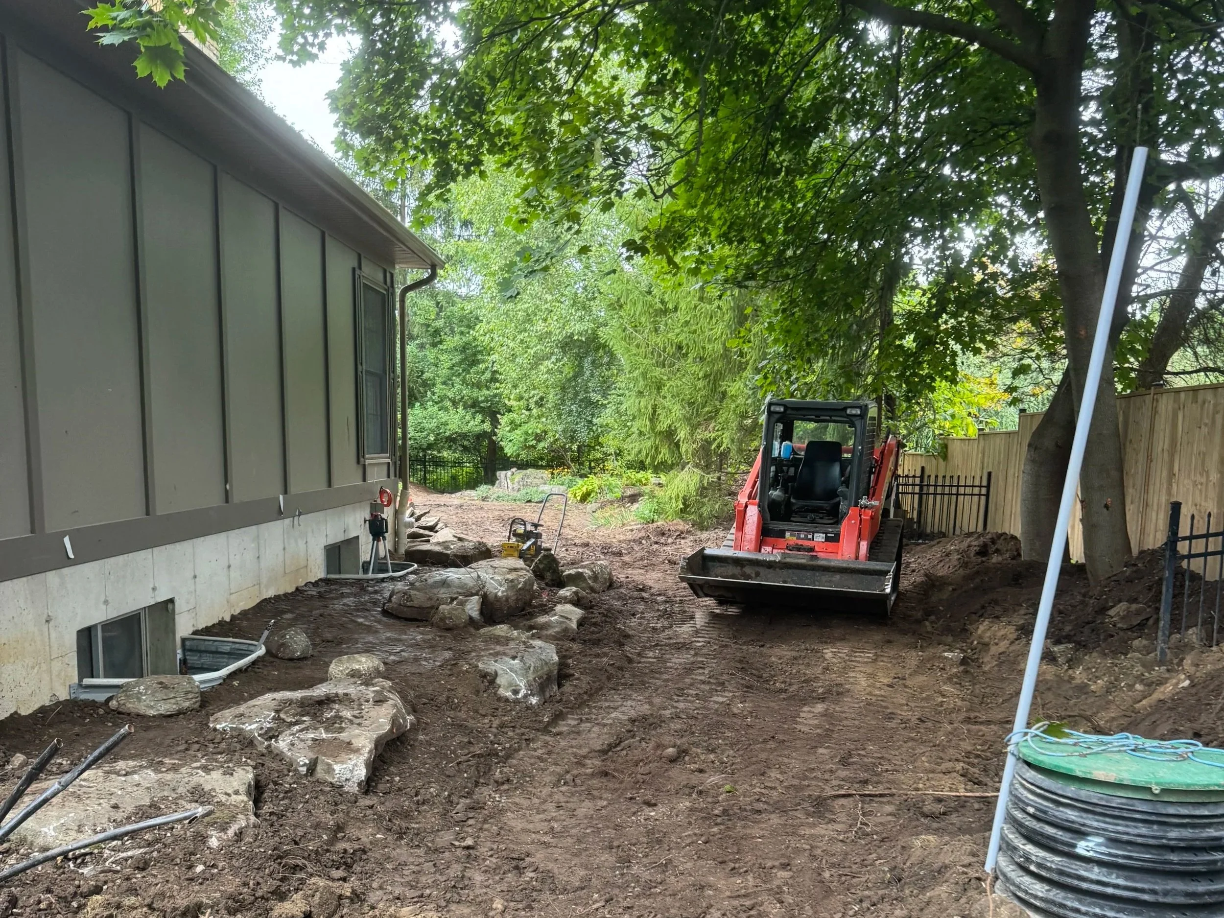 Construction site with a small excavator working near a house, rocks along the foundation, dirt cleared, trees overhead, and a wooden fence in the background.