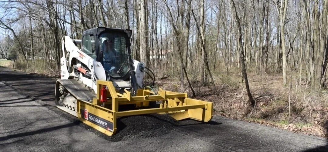 A skid steer loader leveling a section of road for paving amid a wooded area.