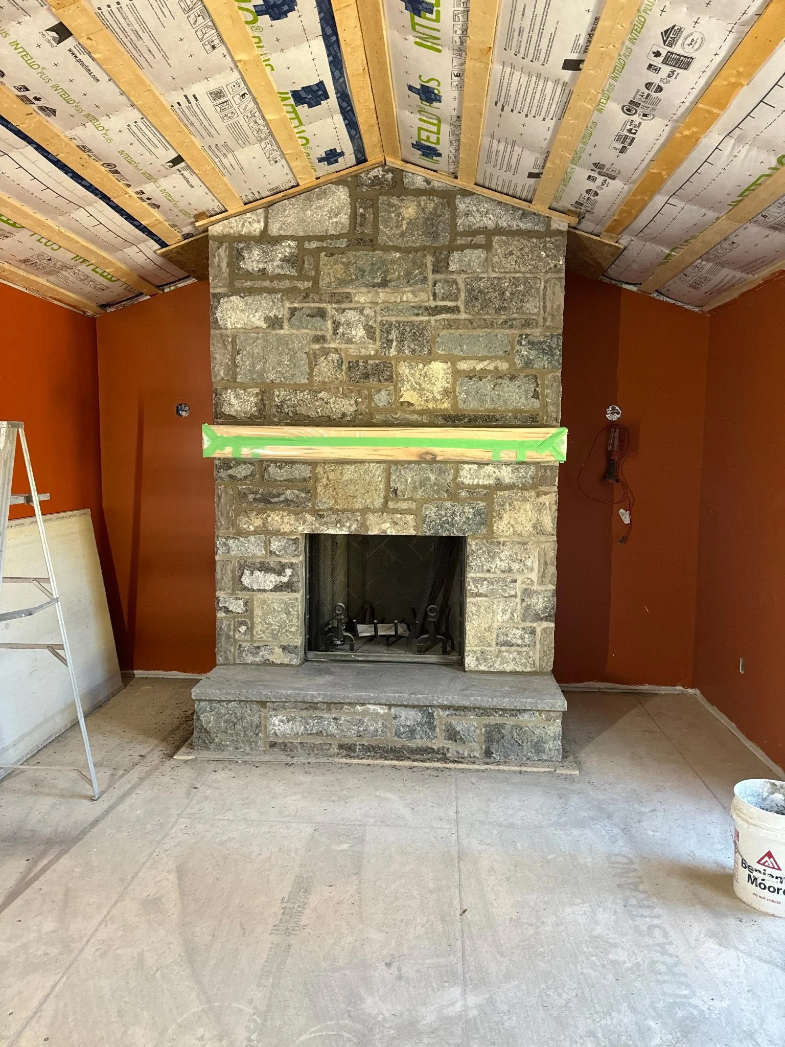 Interior of a room under construction with a stone fireplace, orange walls, and a sloped ceiling covered in insulation material, with construction tools and materials present.