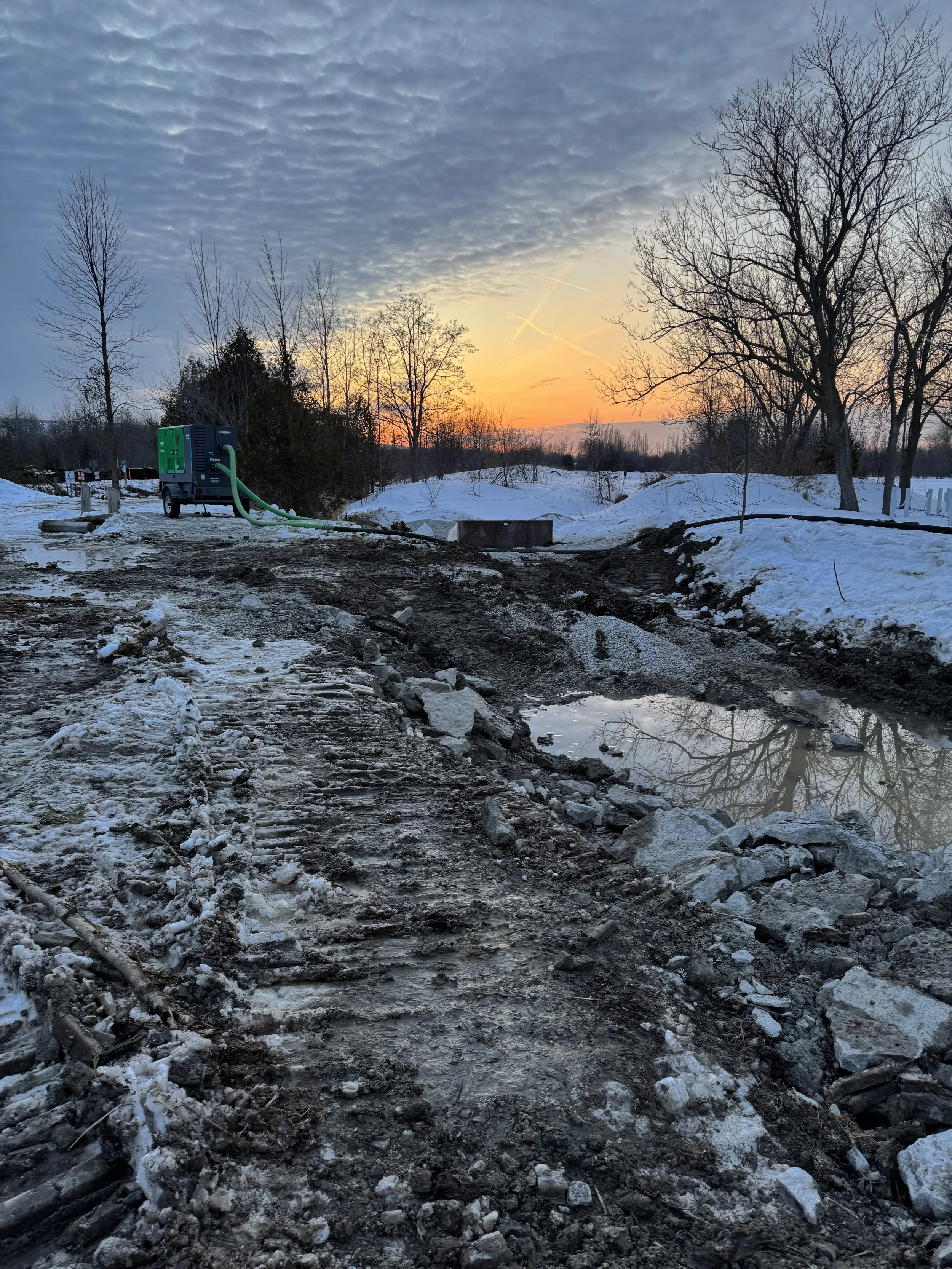 Snowy outdoor scene at sunset with a muddy dirt road, leafless trees, and a blue and green utility machine with a hose.