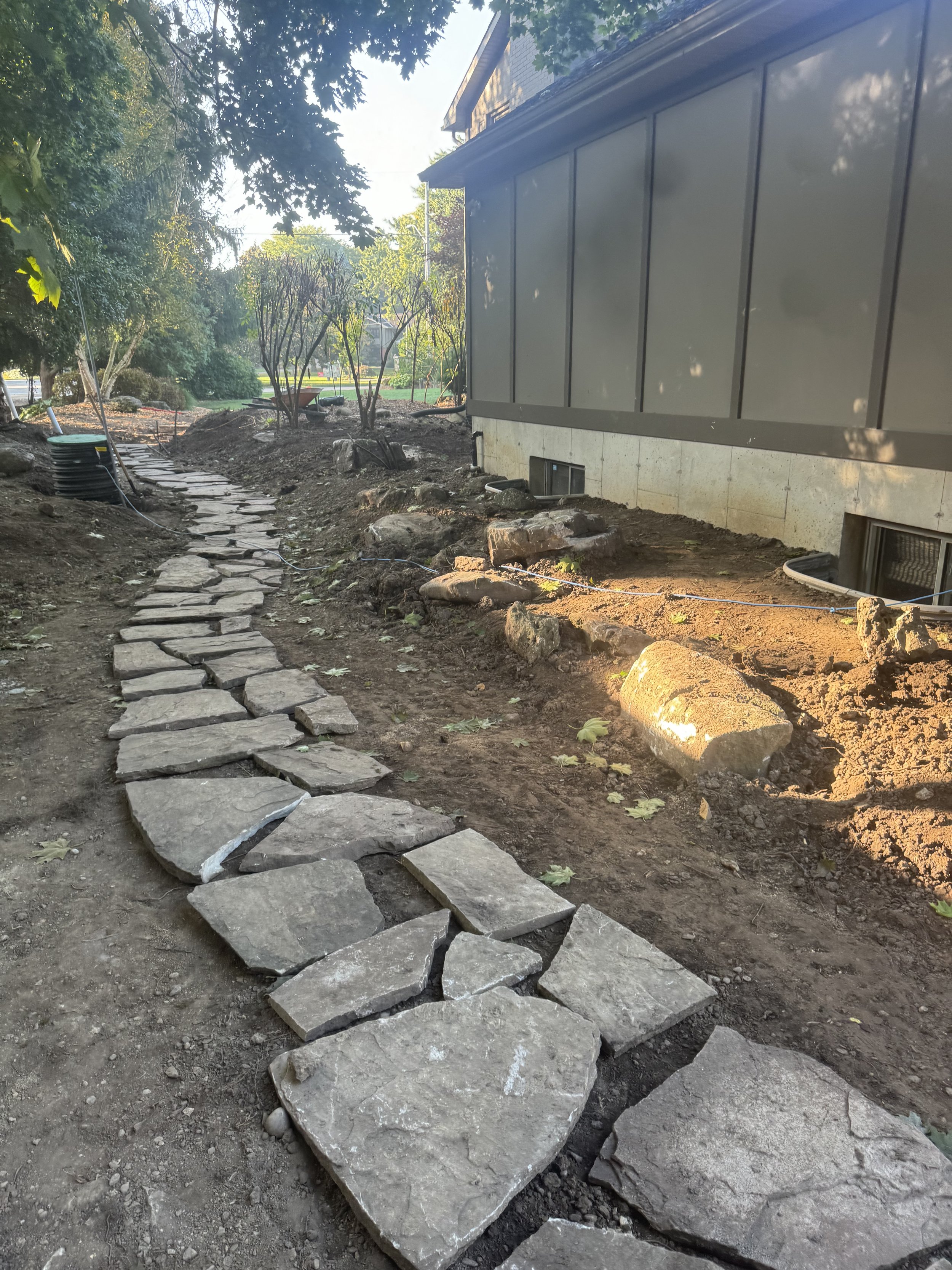 A stone pathway under construction along the side of a building with trees and bushes in the background.
