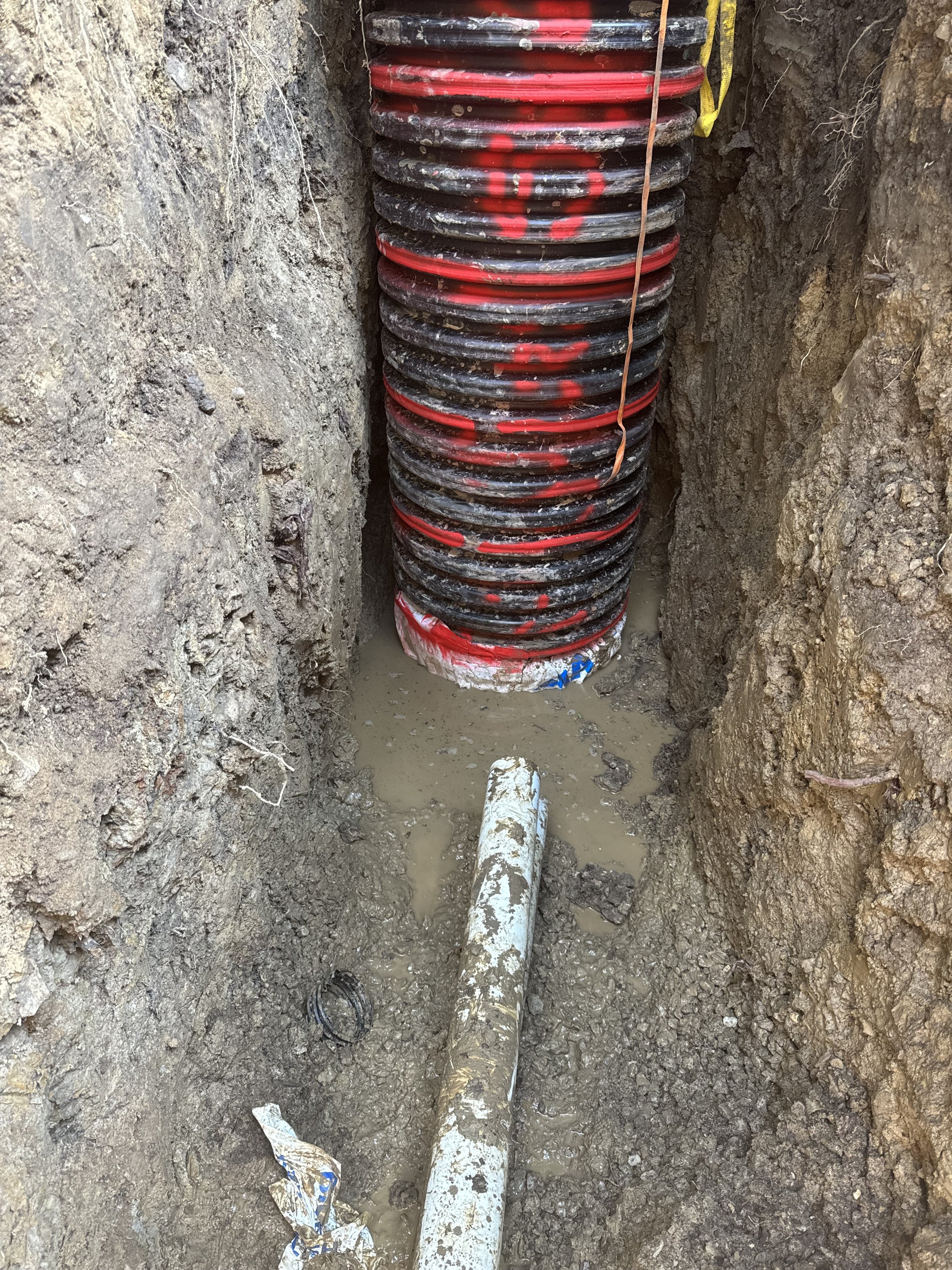 Underground construction site with a piled section of black and red corrugated drainage pipe, partially buried in muddy soil with water pooling around it.