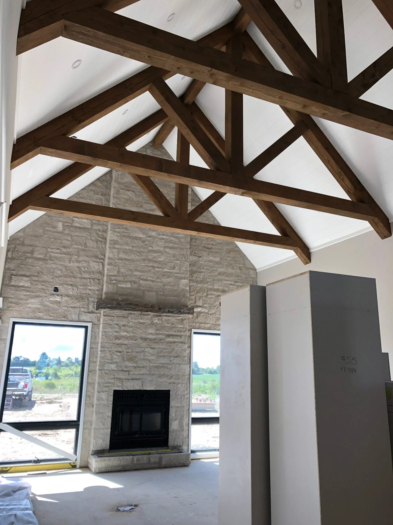 Interior of a house under construction with a vaulted ceiling featuring exposed wooden beams, a stone fireplace, and large windows showing a green landscape outside.