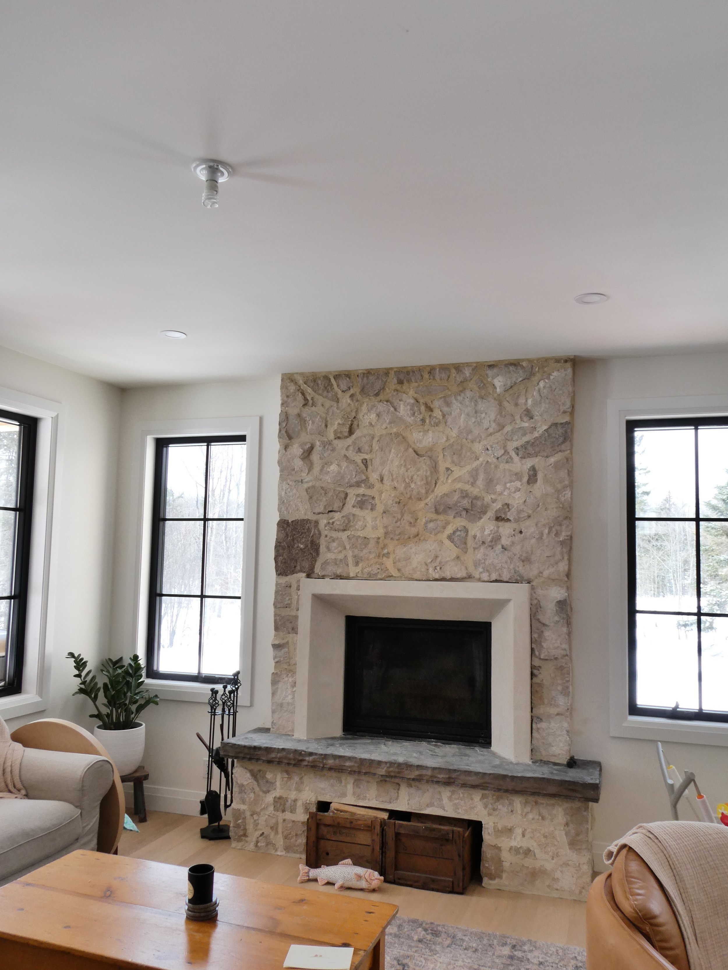 Living room with a stone fireplace, two windows, a beige armchair, a coffee table, and a potted plant.