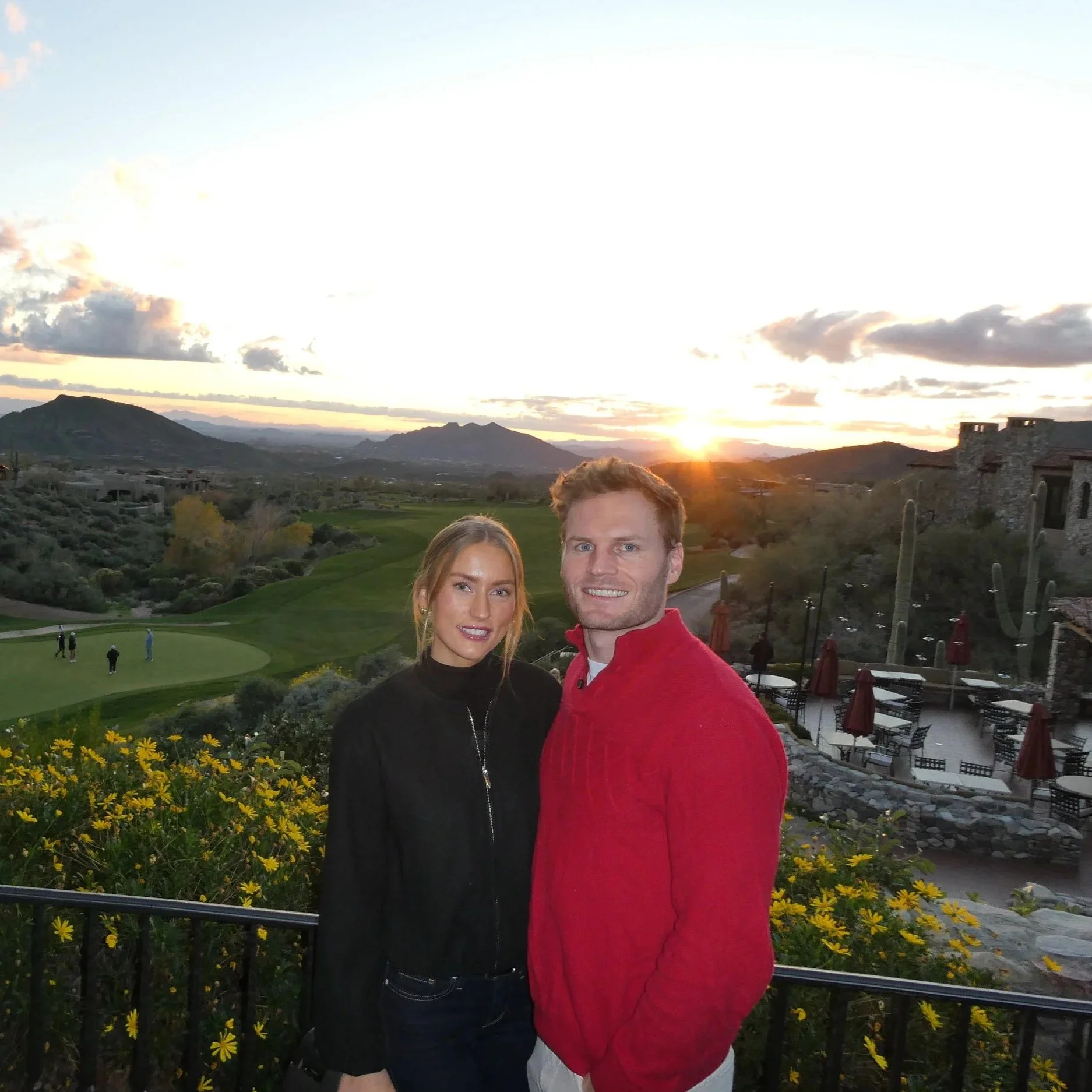 A smiling couple standing outdoors during sunset, with a golf course, mountains, and desert landscape in the background.