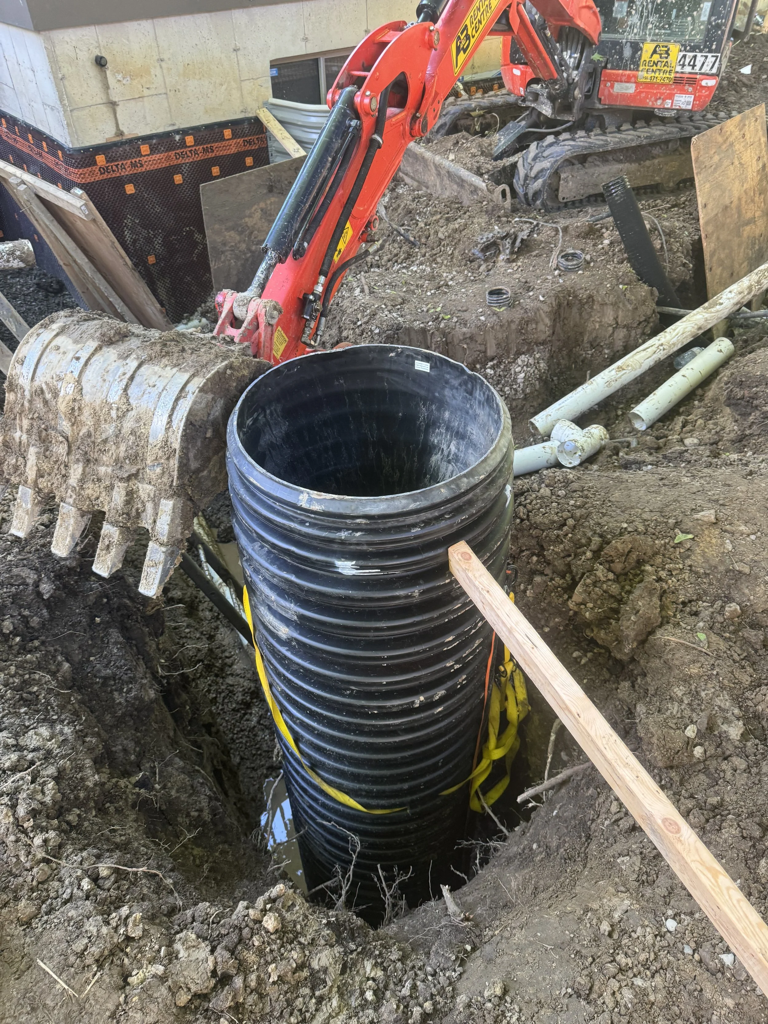 Excavation site with black corrugated pipe being lowered into a trench, a mini excavator with a bucket attachment, dirt, and construction pipes nearby.Exterior Sumpbasin