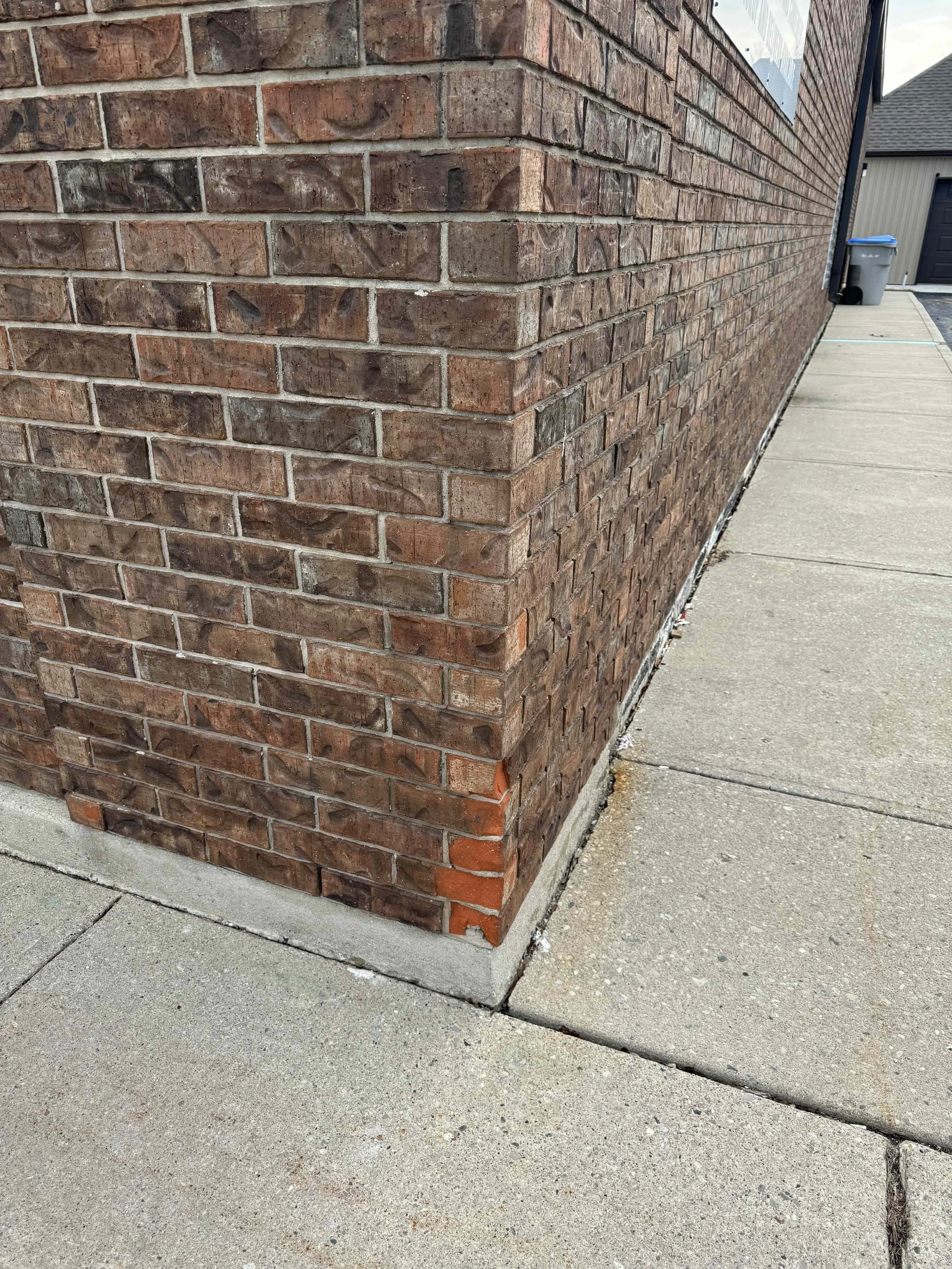 Close-up view of the corner of a brick house with a concrete sidewalk nearby, showing some damaged brickwork at the base.