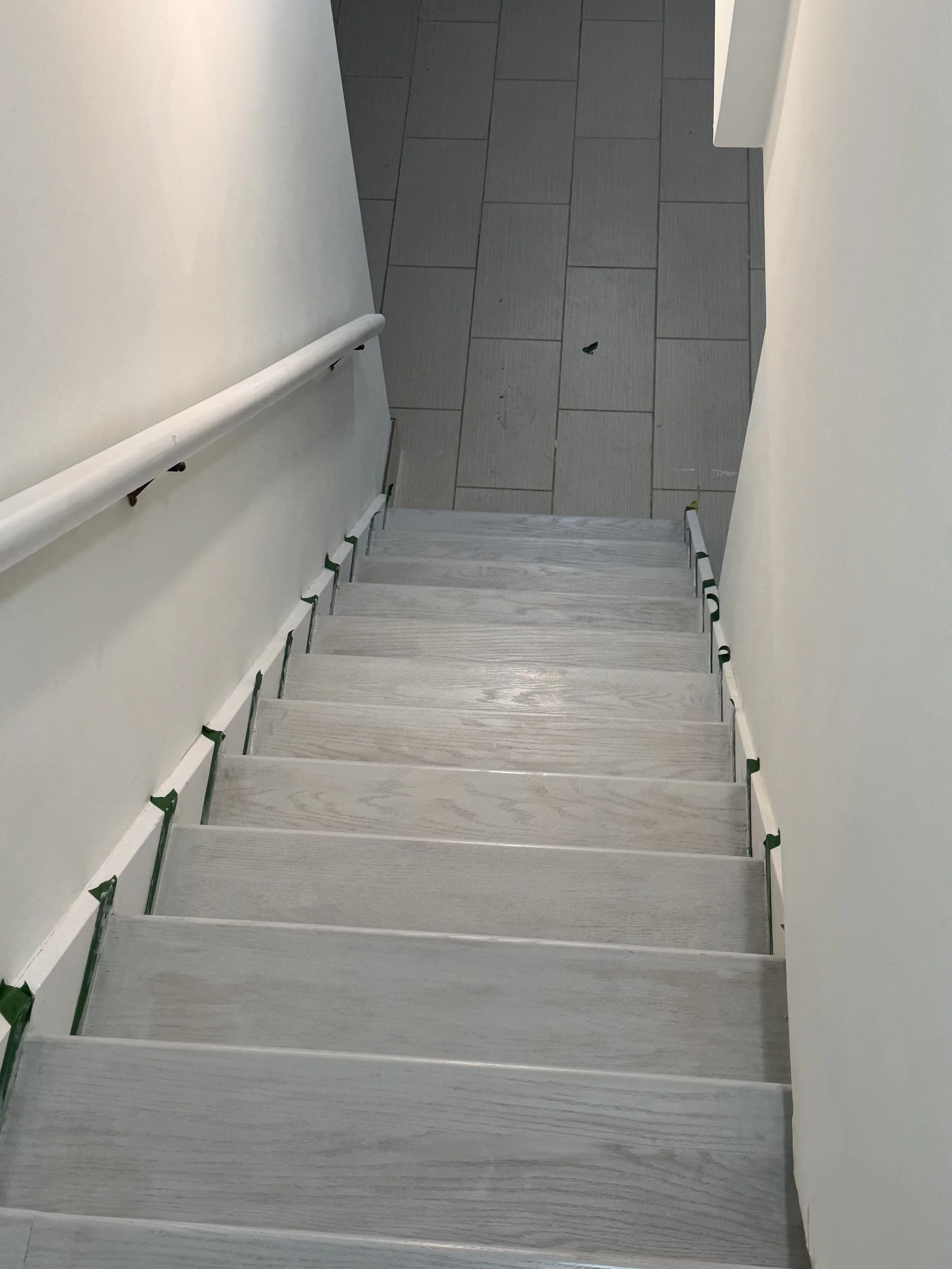 View of a staircase from above, with light gray wooden steps, a white handrail on the left, and gray tile flooring at the top of the stairs.