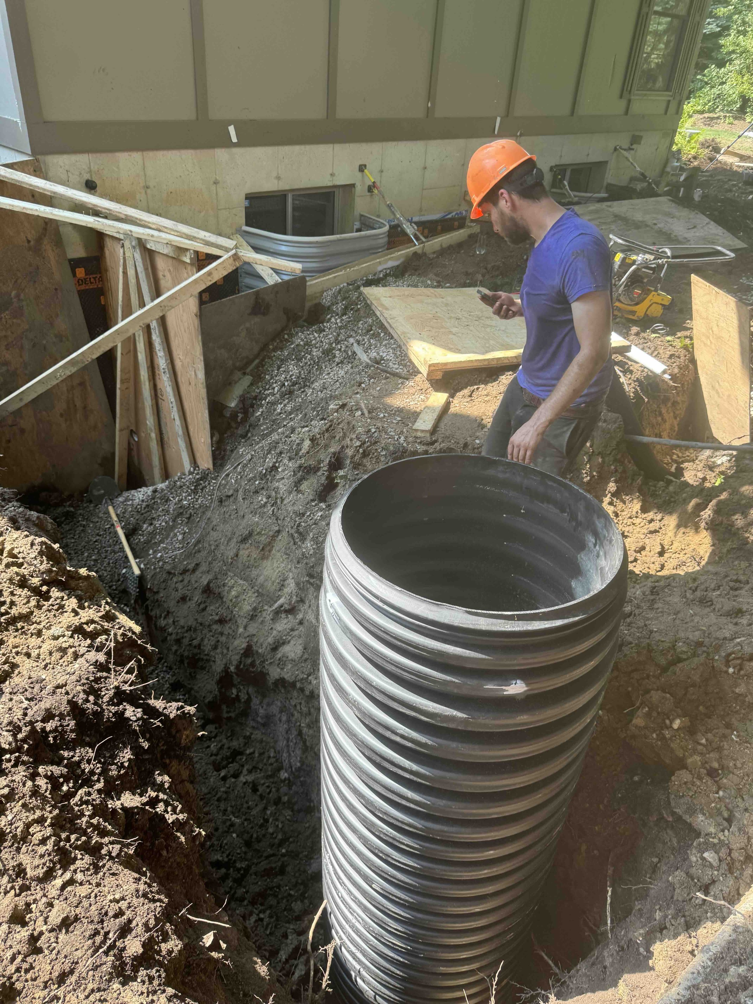 A construction worker with an orange safety helmet and blue shirt standing in a dug site, looking at his phone with a large black pipe beside him and construction materials and tools around.