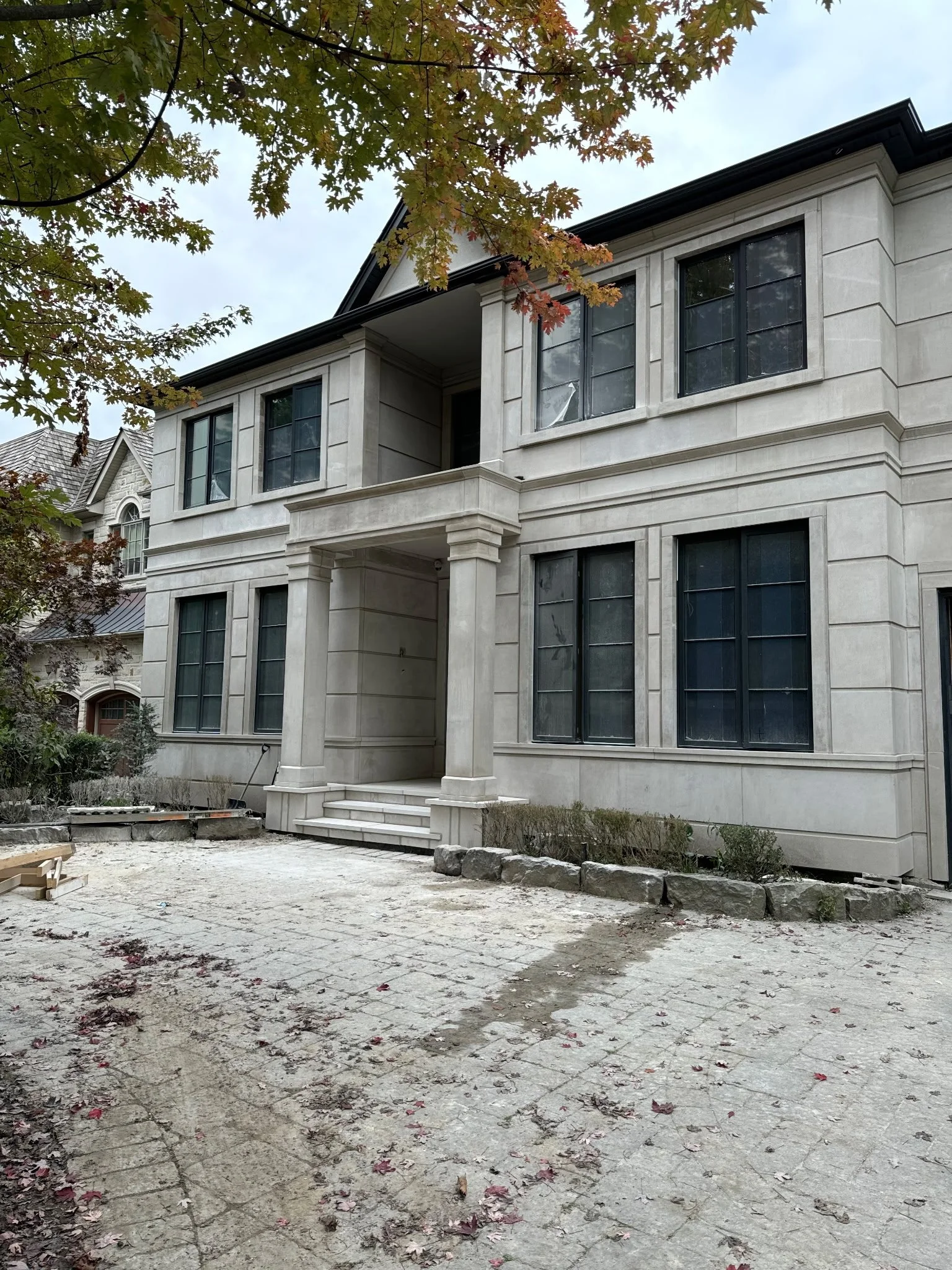 Front of a multi-story house under construction with large windows, a small front staircase, and a dirt driveway, surrounded by trees with autumn leaves.