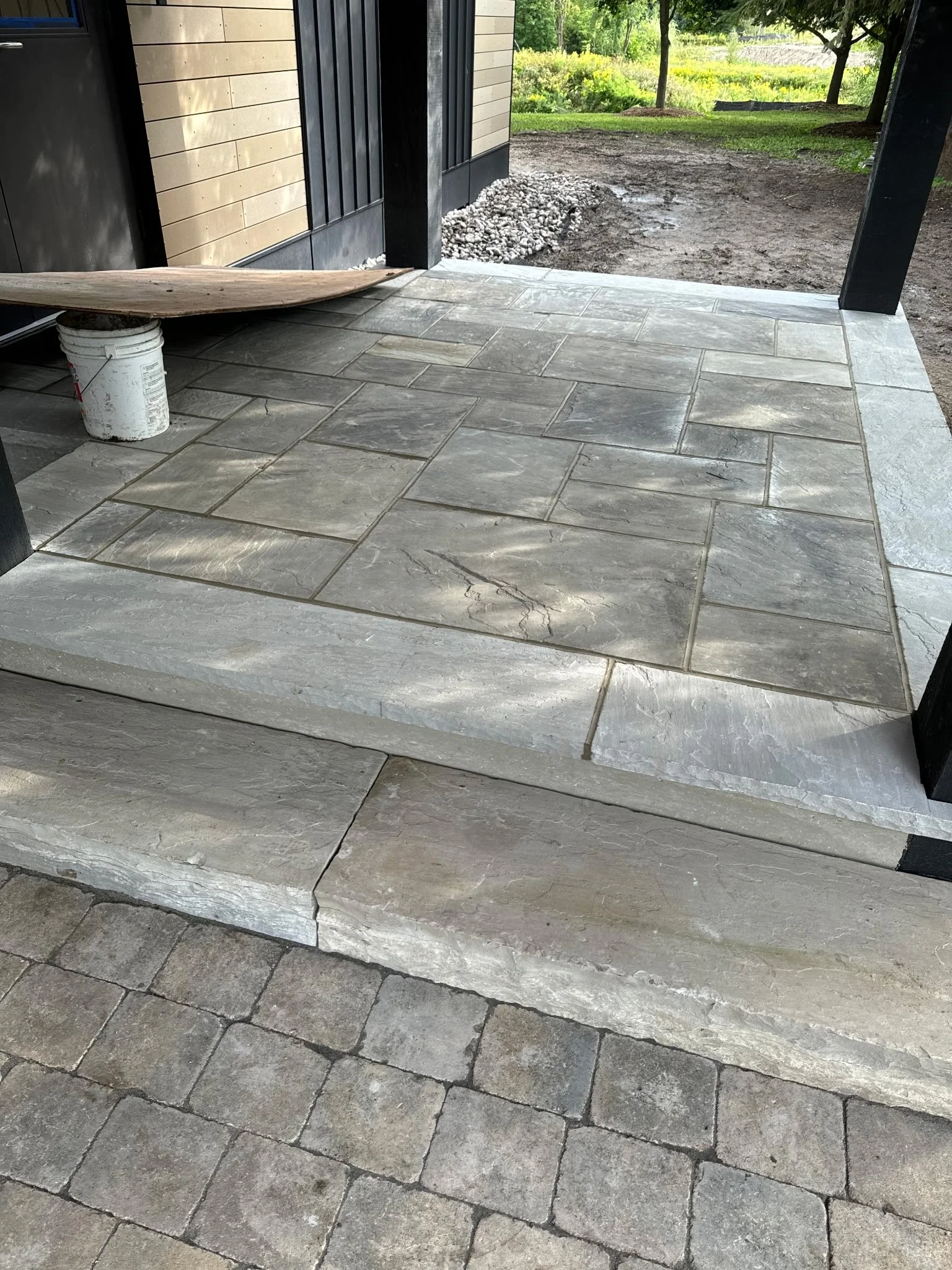 View of a porch with new stone tiles, a step with a crack, an outdoor landscape with trees and bushes, and a dirt pathway.