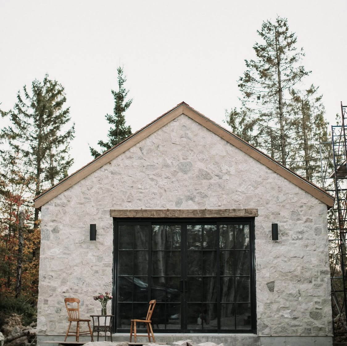 A stone house with large black framed glass doors, two chairs, a small table with a flower vase outside, surrounded by tall trees.