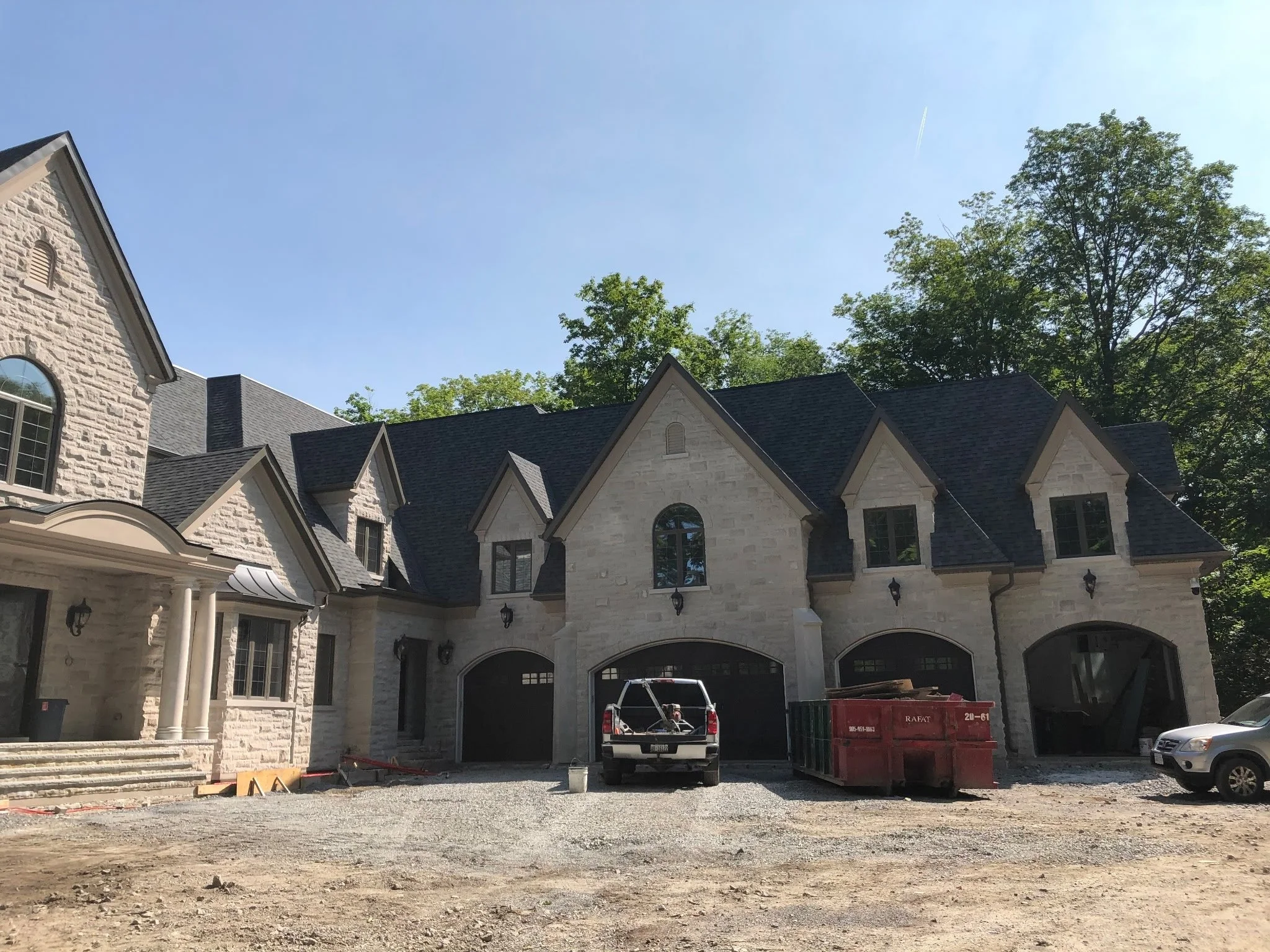 Large house under construction with a gravel driveway, a pickup truck, a red dumpster, and a silver car parked outside. The house has multiple gabled roofs, arched windows, and stone exterior walls, with some construction materials visible.
