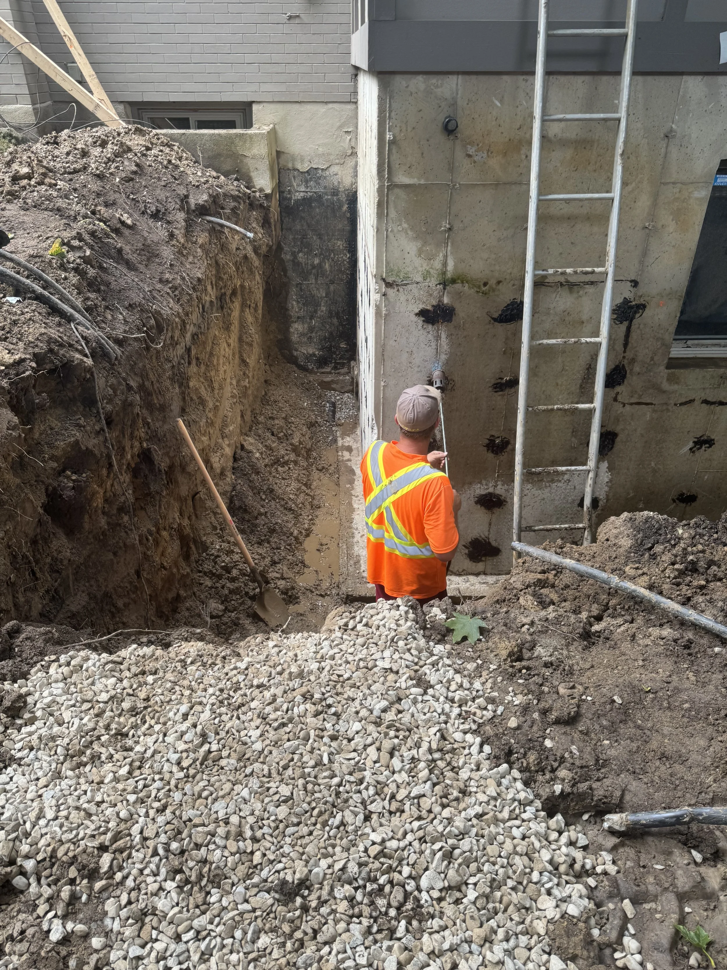 Construction worker in bright orange safety vest and hat inspecting a concrete foundation wall with a ladder against it, surrounded by dirt and gravel.