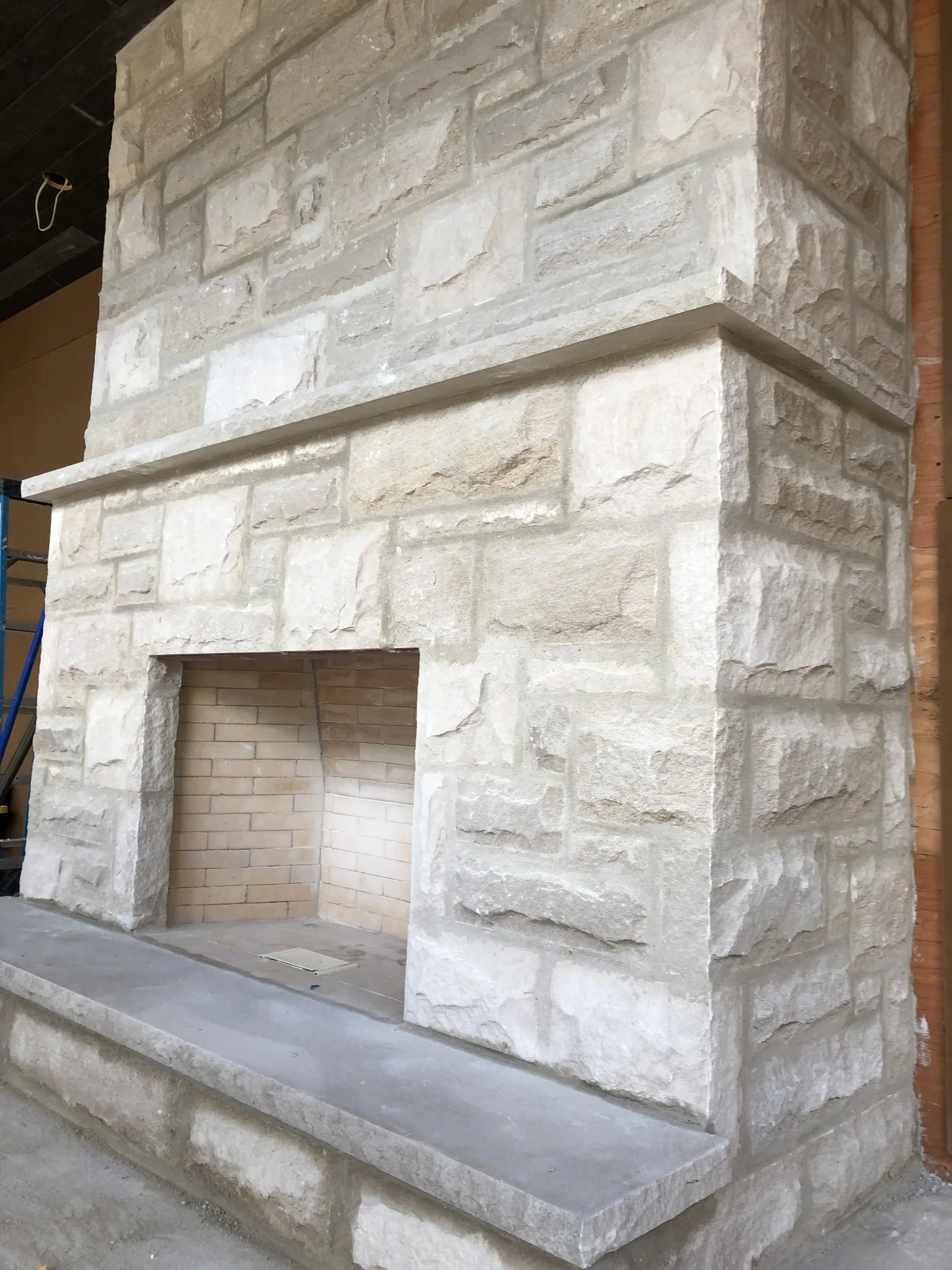 Close-up view of a stone fireplace under construction, showing the exterior facade with large light-colored stones and an open interior lined with small brick tiles.