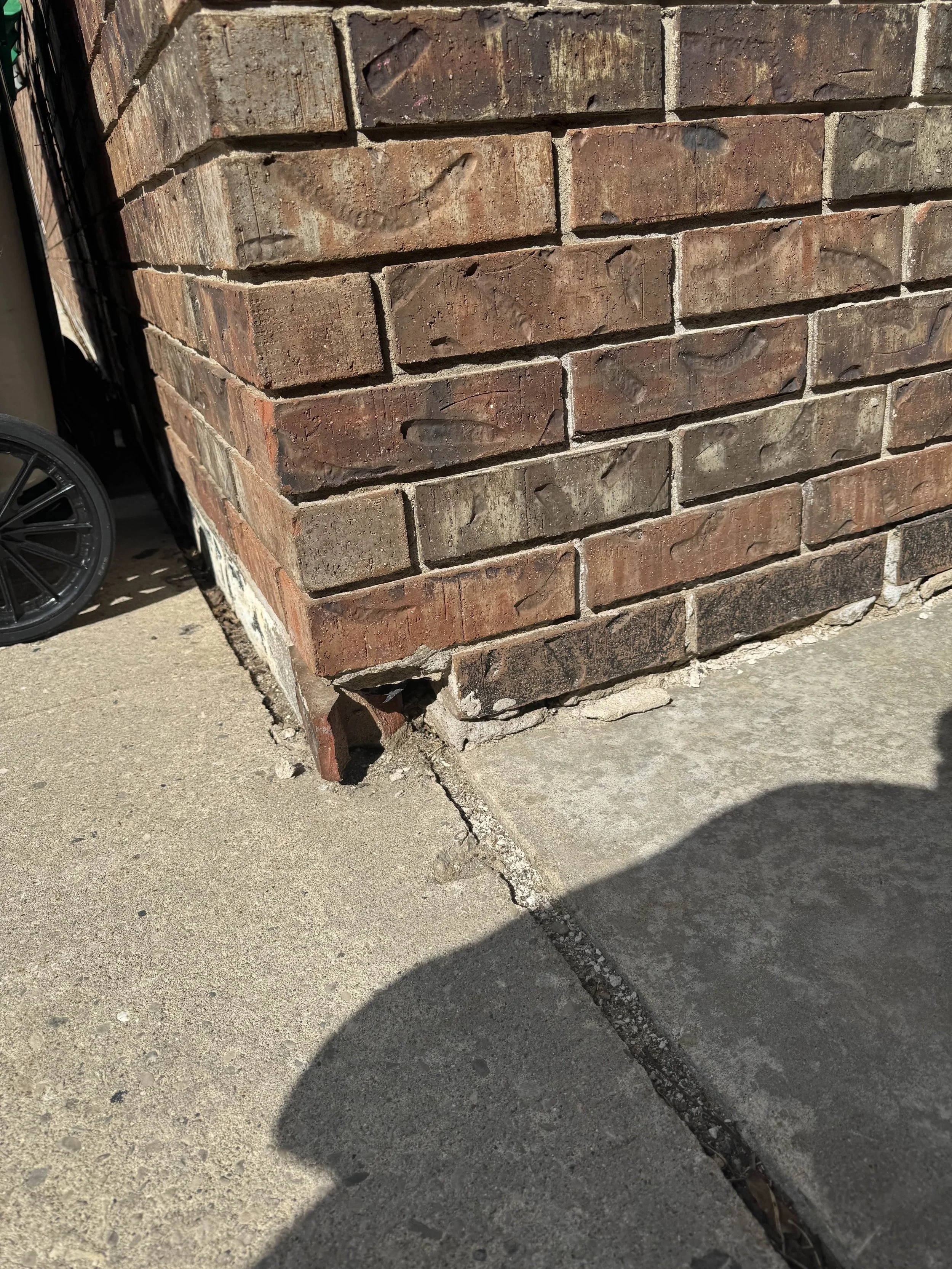 Corner of a brick wall with visible damage at the bottom, where a brick is missing. The wall meets a concrete sidewalk. Shadows are cast on the sidewalk, indicating sunlight.