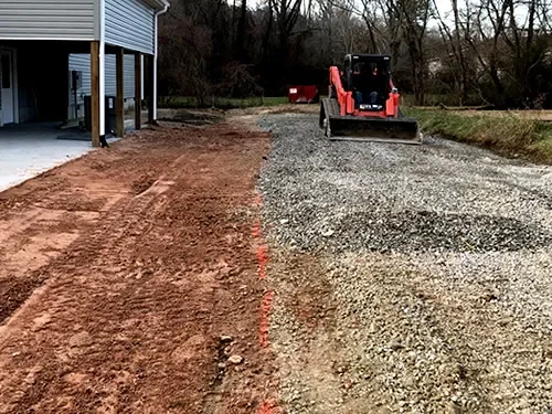 A construction site with a partially gravelled driveway, a small red compact loader, and a building on the left.