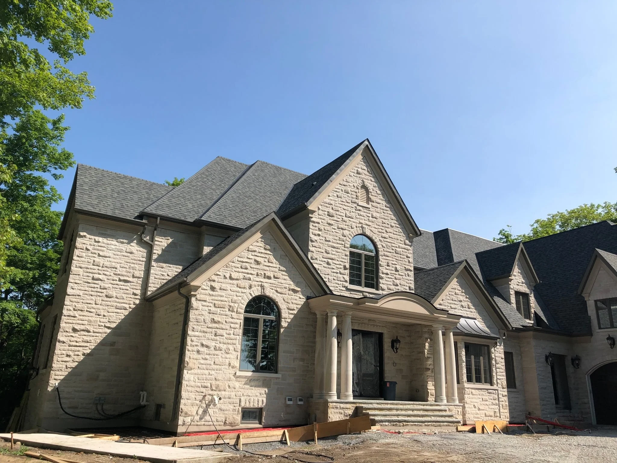 A large, elegant two-story house with light-colored stone exterior walls, arched and rectangular windows, a peaked roof, and a front porch with columns, under a clear blue sky.