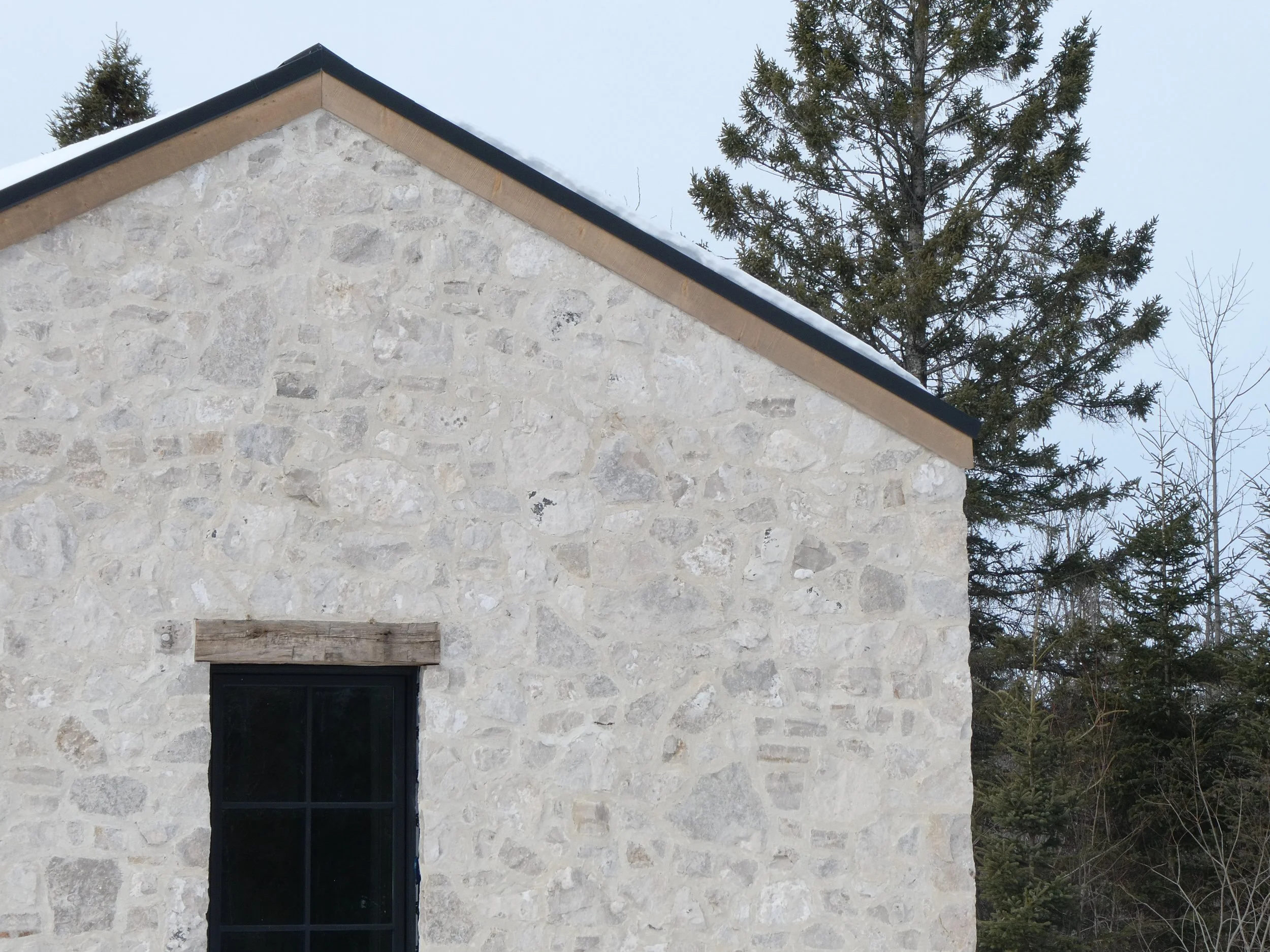 Close-up of a stone house wall with a black-framed window, a wooden trim above the window, and a snow-covered roof, with pine trees in the background.