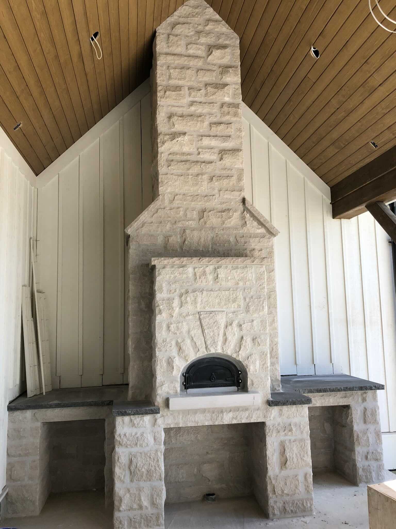 Stone outdoor fireplace with black oven door, built-in counters on each side, in a room with wood-paneled ceiling and white paneled walls.