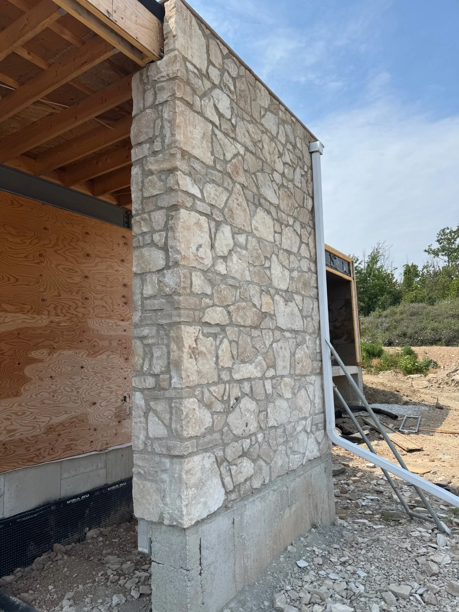 Construction site with a stone and concrete wall, wooden framing, and exposed piping under a blue sky.