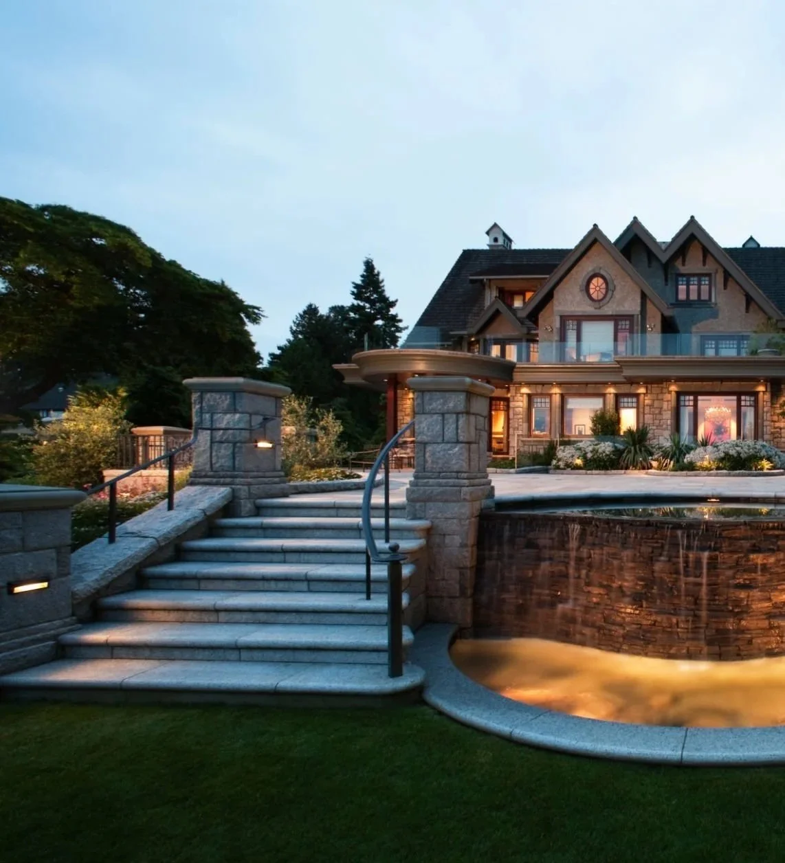 A luxurious house with stone steps leading to a water feature. The house has a large porch, multiple dormer windows, and warm interior lighting, surrounded by landscaped gardens at dusk.