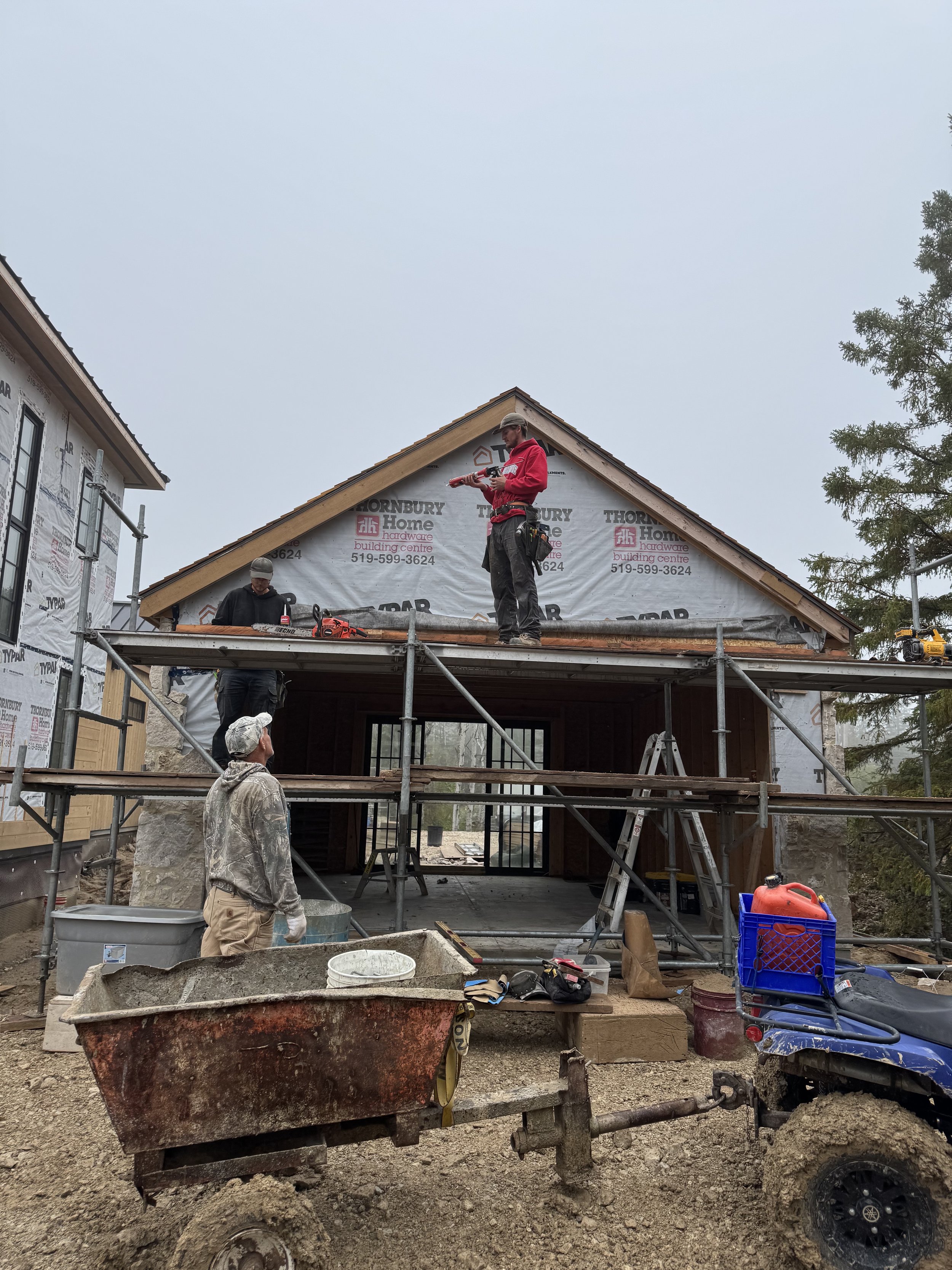 Construction workers working on a house frame, with scaffolding, tools, and construction materials around, during daytime.