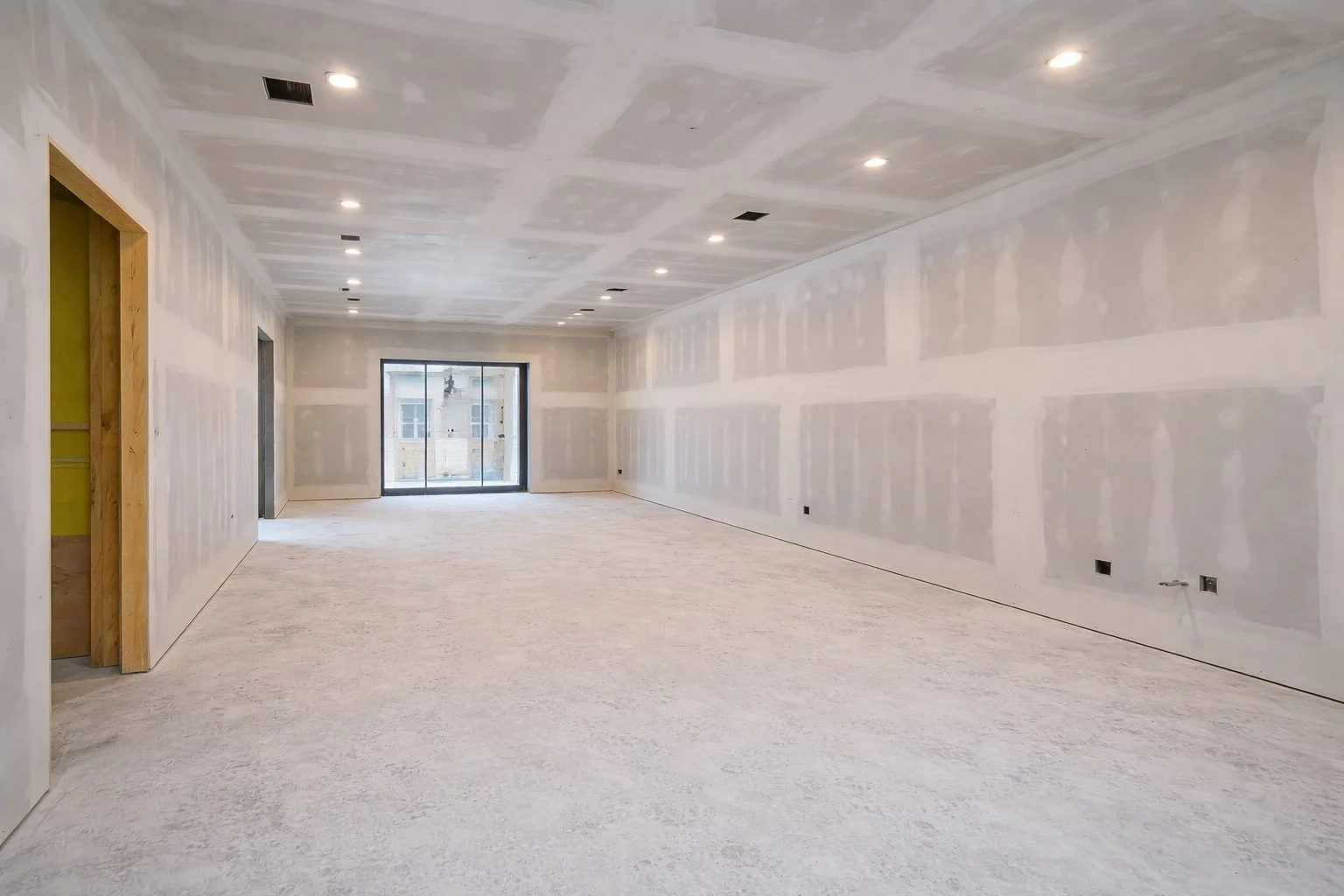 Empty room under construction with unfinished drywall on walls and ceiling, recessed lighting installed, sliding glass door at the far end, and floors covered with protective material.