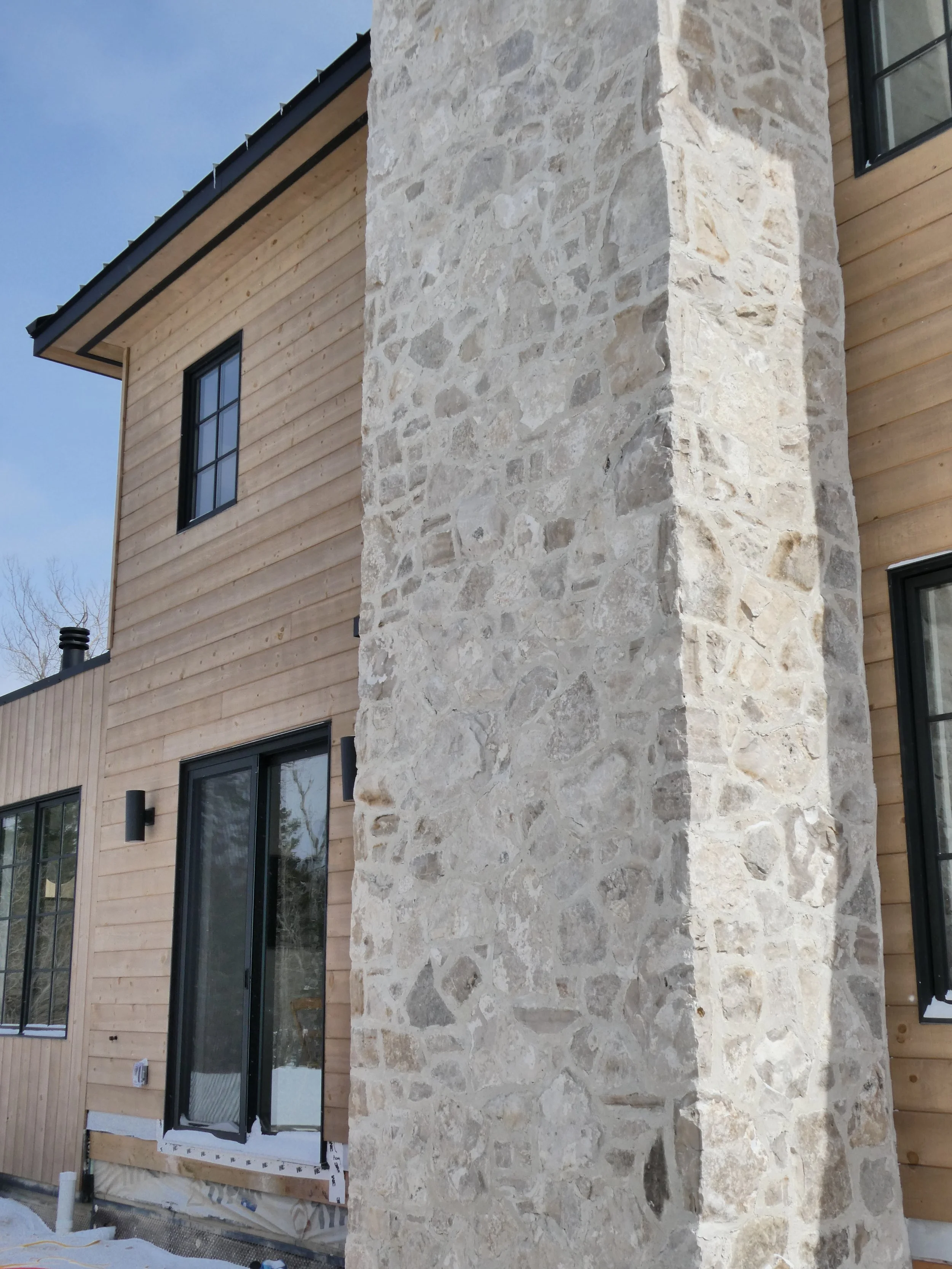 Close-up of a modern house exterior showing a large stone chimney, wooden siding, black-framed windows, and a sliding glass door.