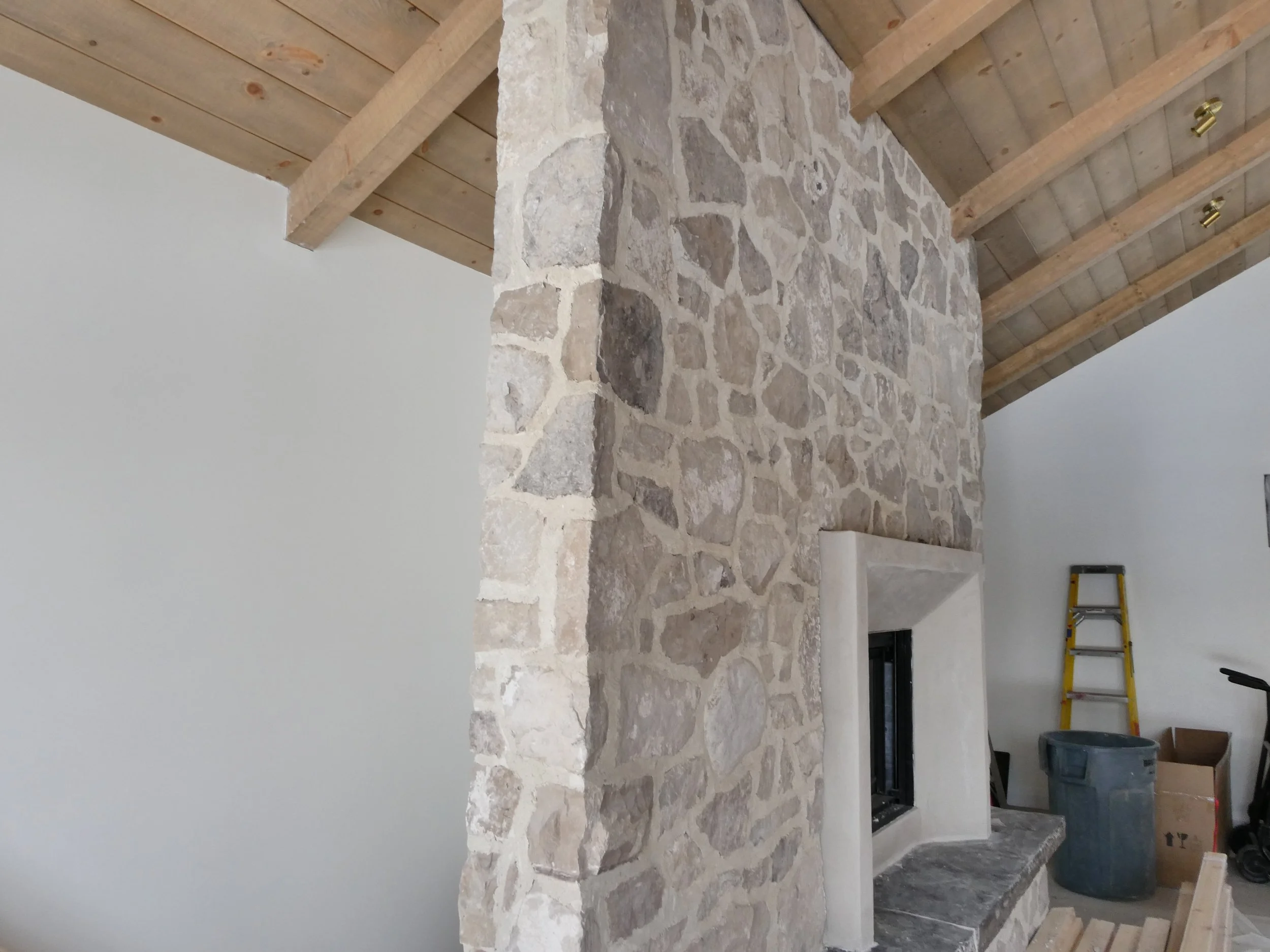 Interior view of a room under construction with a stone fireplace, unfinished wooden ceiling, ladder, trash can, and boxes.