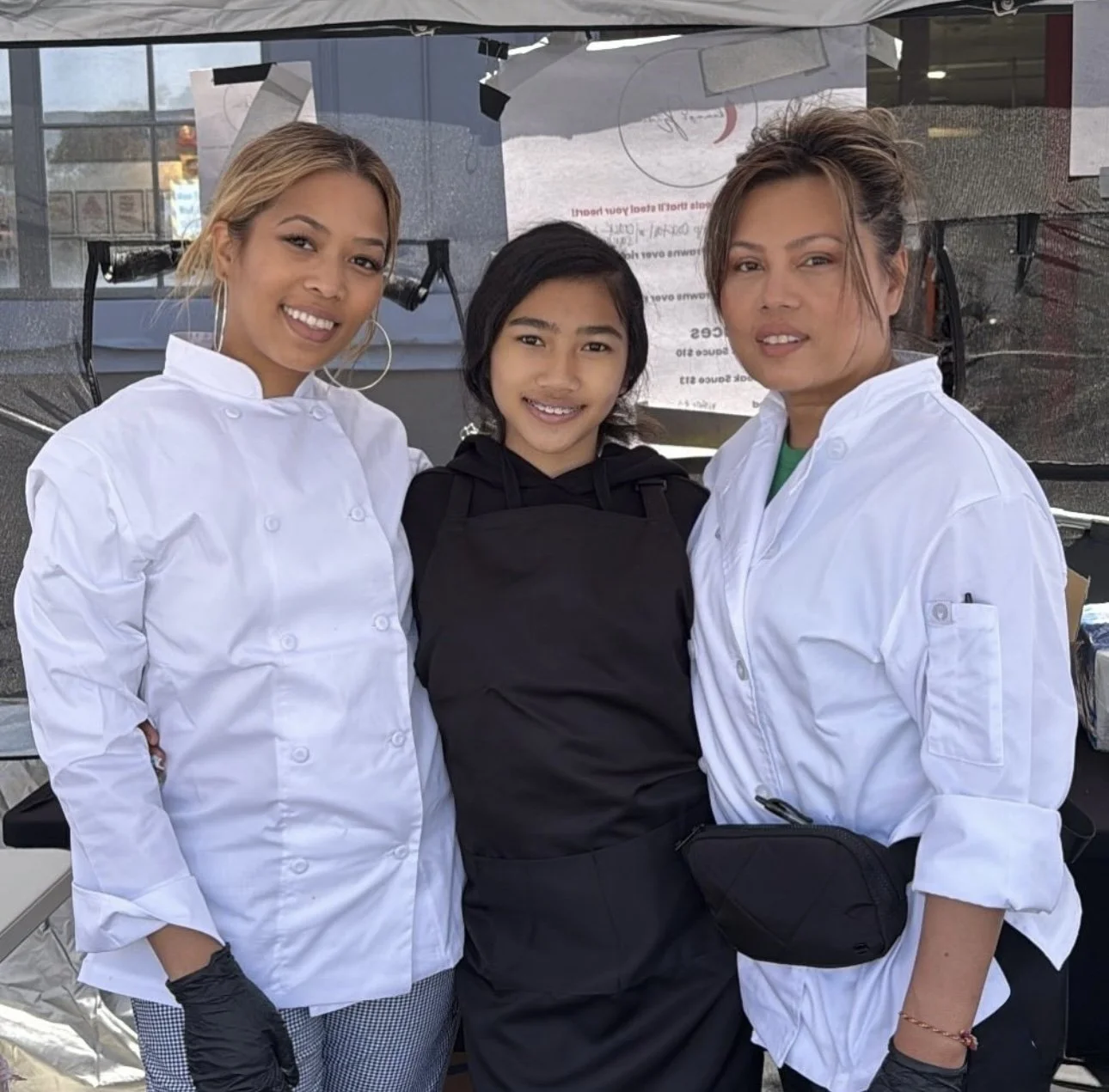 Three women, two wearing chef coats and one in a black hoodie, standing close together and smiling outdoors.