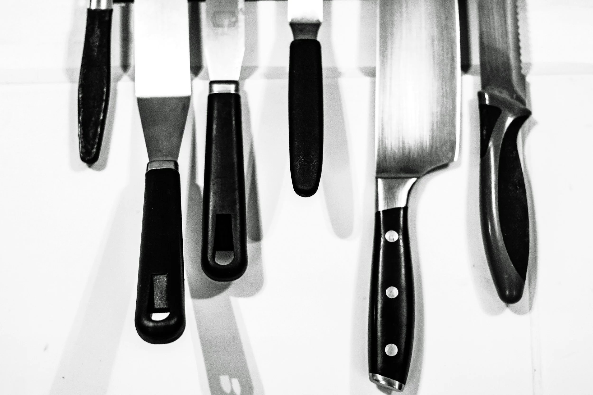 A collection of kitchen knives with black handles hanging on a wall. The knives vary in size and shape, and one knife has a serrated edge.