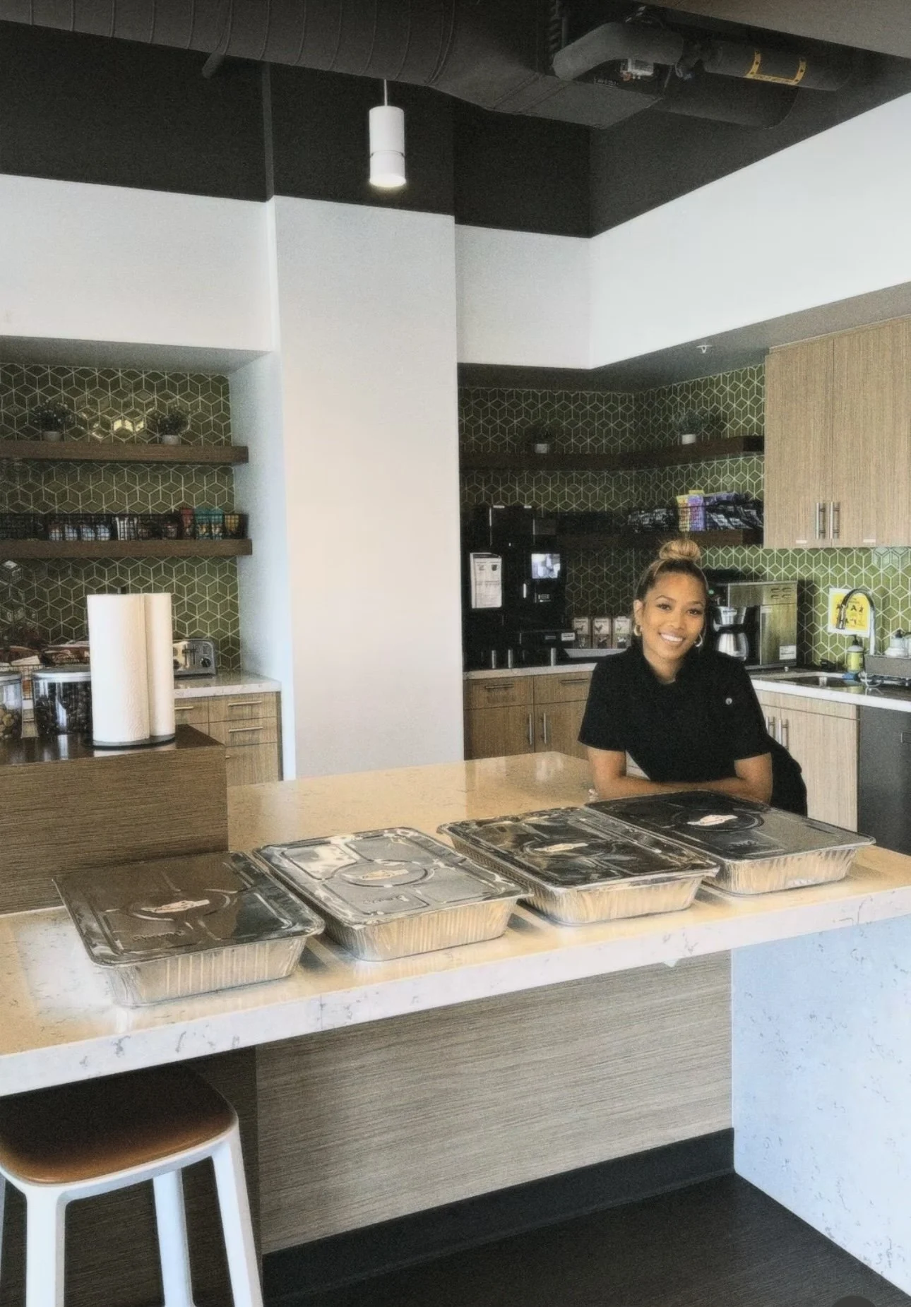 A woman standing behind a kitchen counter with four foil-covered containers, smiling at the camera.