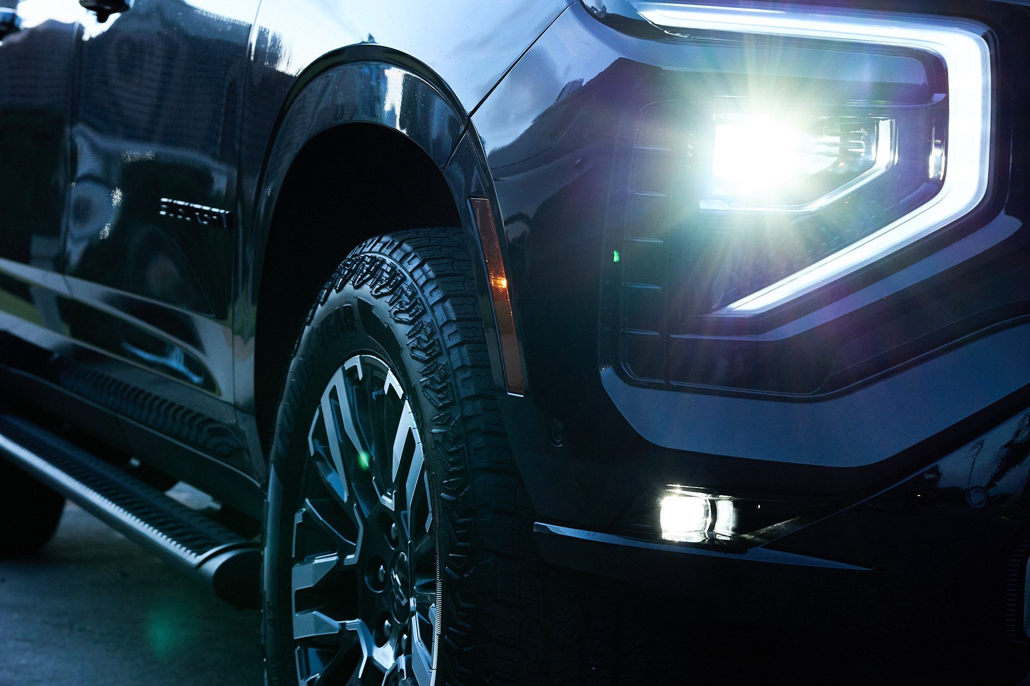 Close-up of the front passenger side of a black pickup truck with a headlight turned on, highlighting the tire, wheel, and body details.
