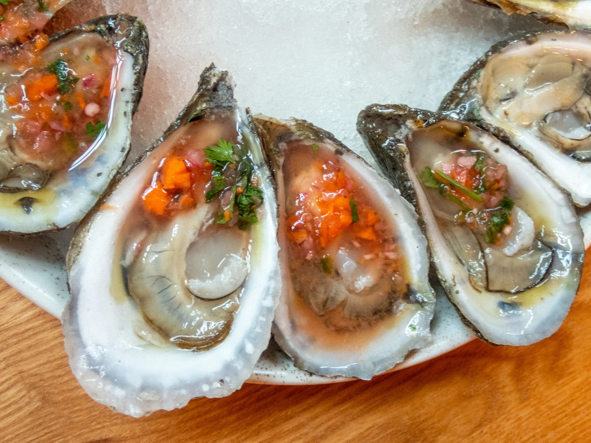 Close-up of raw oysters on a plate, garnished with chopped vegetables and herbs, with a wooden surface underneath.