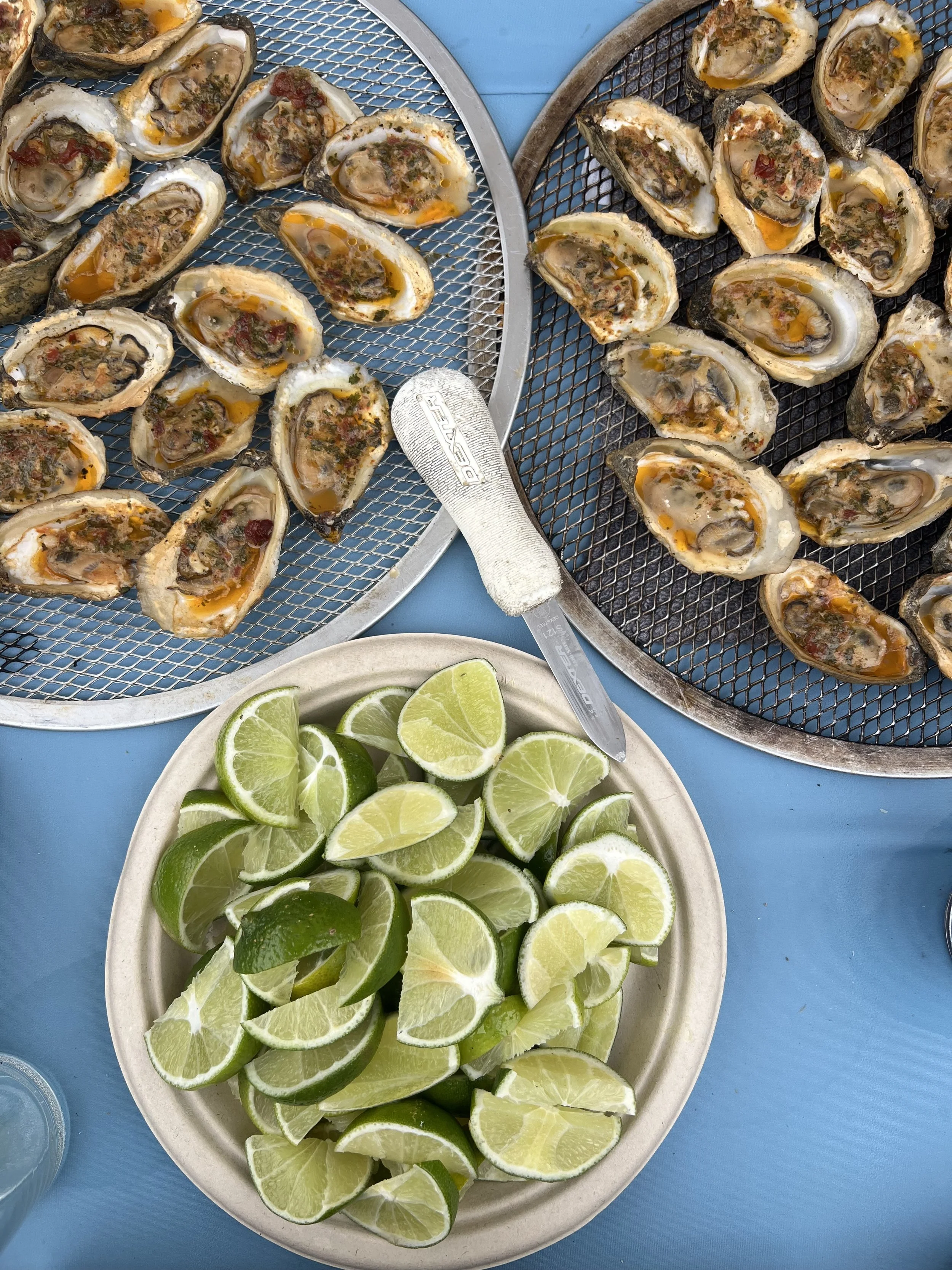 Fresh oysters on a tray with chopped herbs and cheese, with a plate of lime wedges nearby.