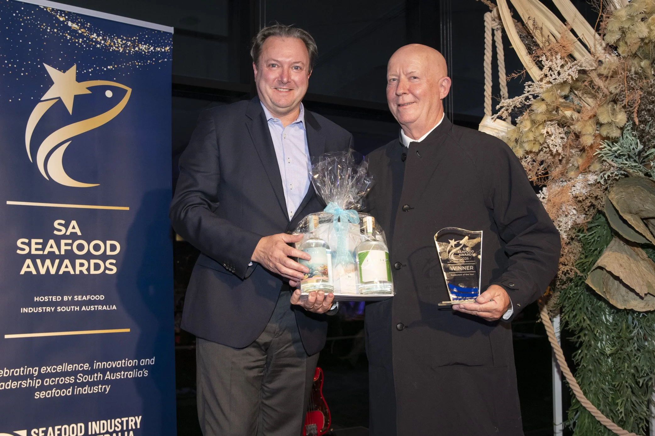 Person in black shirt and striped apron holding a black plate with a cooked lobster and garnishes, at the SA Seafood Awards 2026 event.