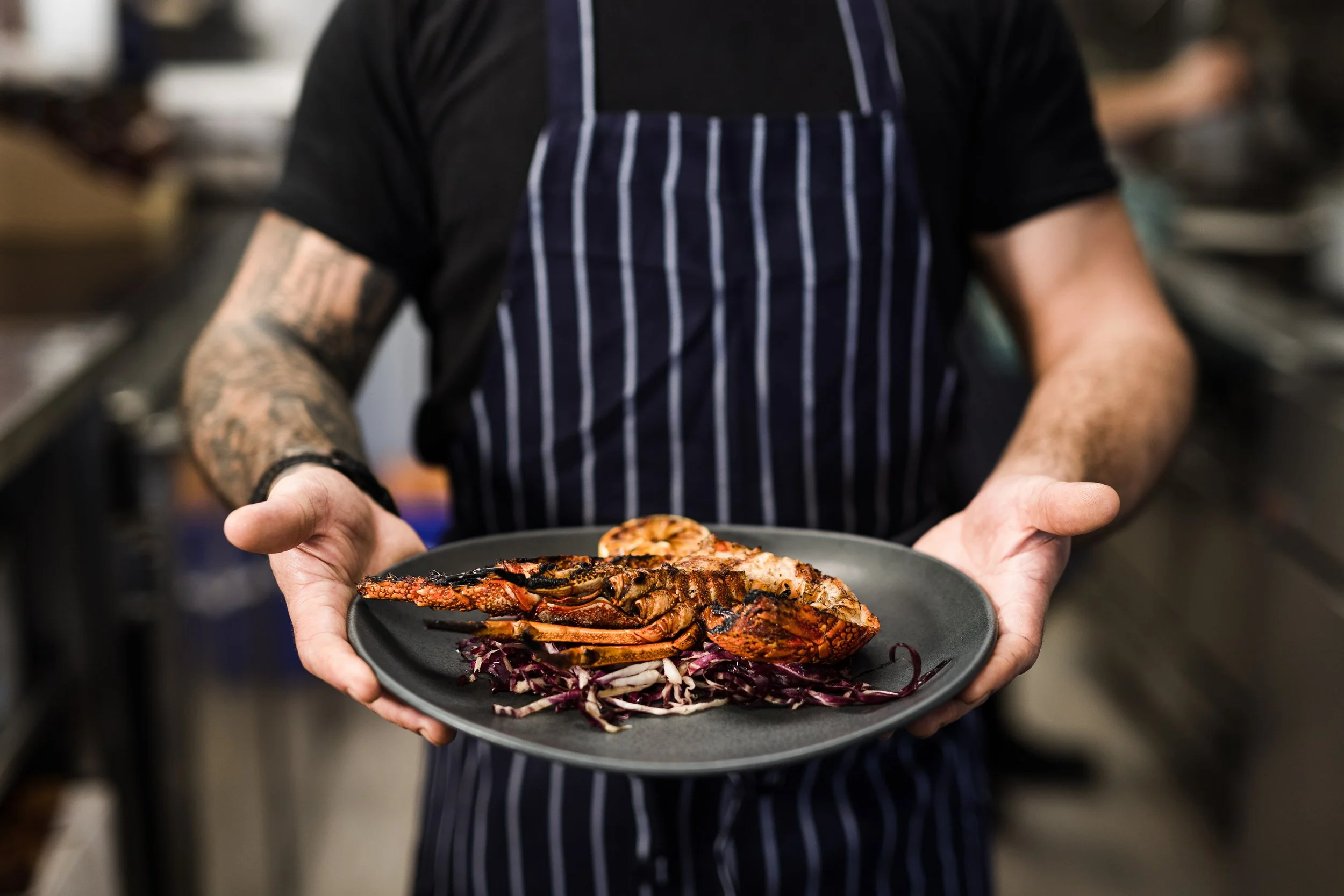 Person in a striped apron holding a black plate with a cooked lobster, garnished with purple cabbage slaw.