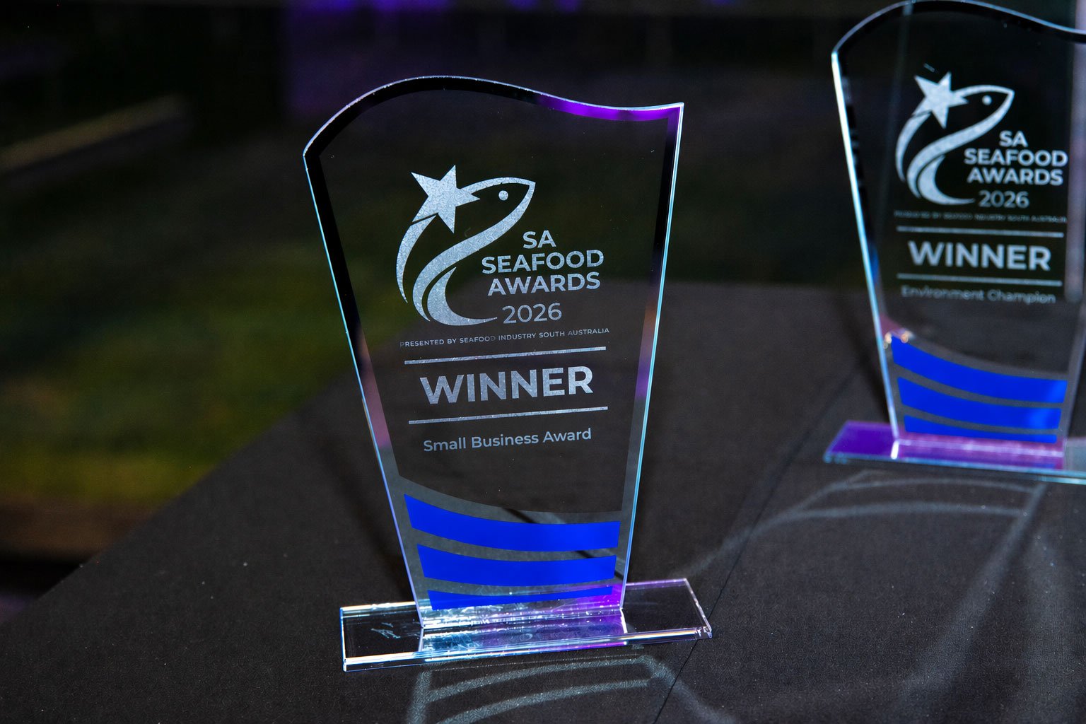 A woman wearing a blue shirt is selecting seafood from a display case at a seafood market. The display includes prices for different types of fish.
