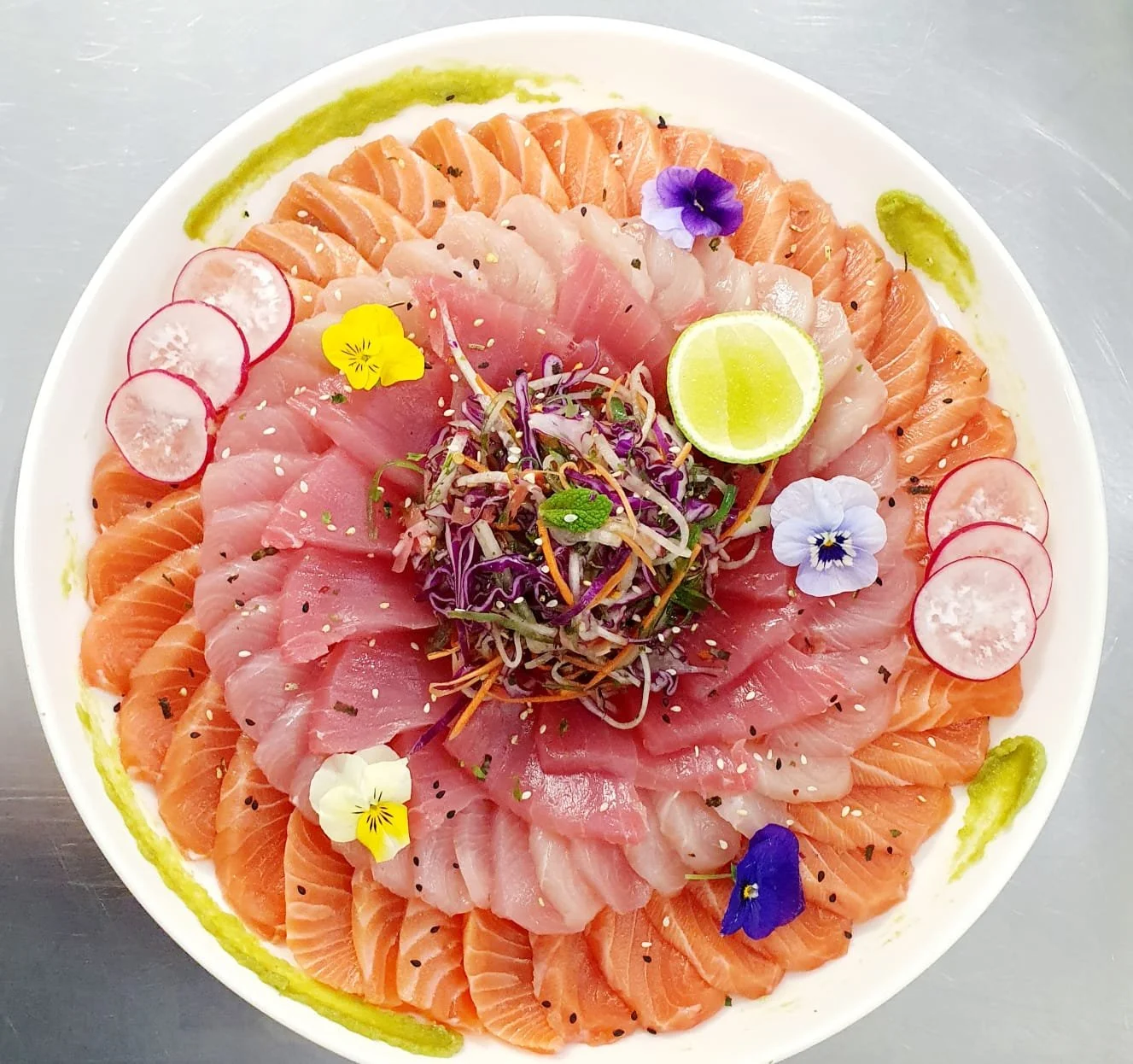 Assorted sashimi, including kingfish and tuna, arranged in a circle on a white plate, garnished with radish slices, edible flowers, a lime wedge, and sprinkled with sesame seeds and herbs.
