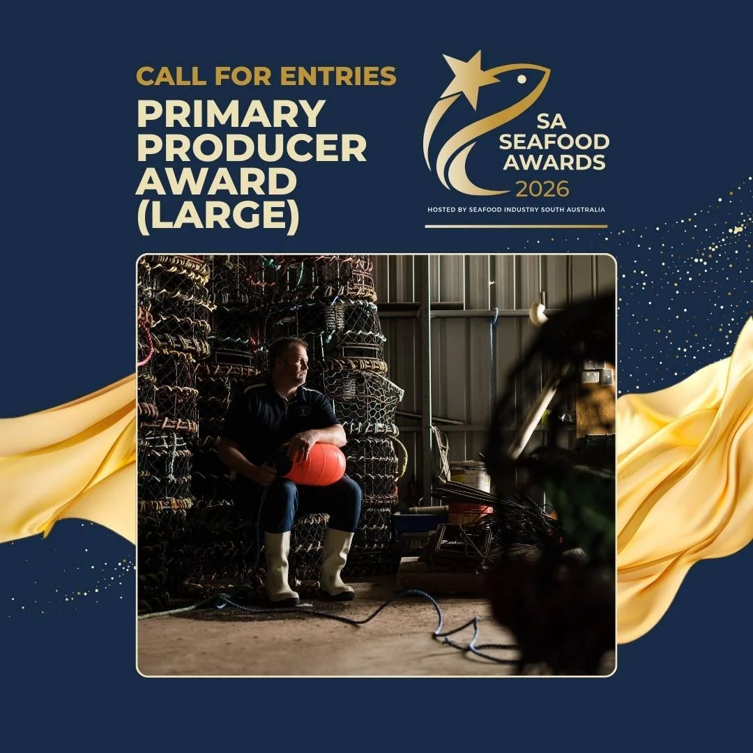 A person sitting on a stack of lobster traps inside a seafood storage warehouse, holding a red buoy, with machinery and equipment around.